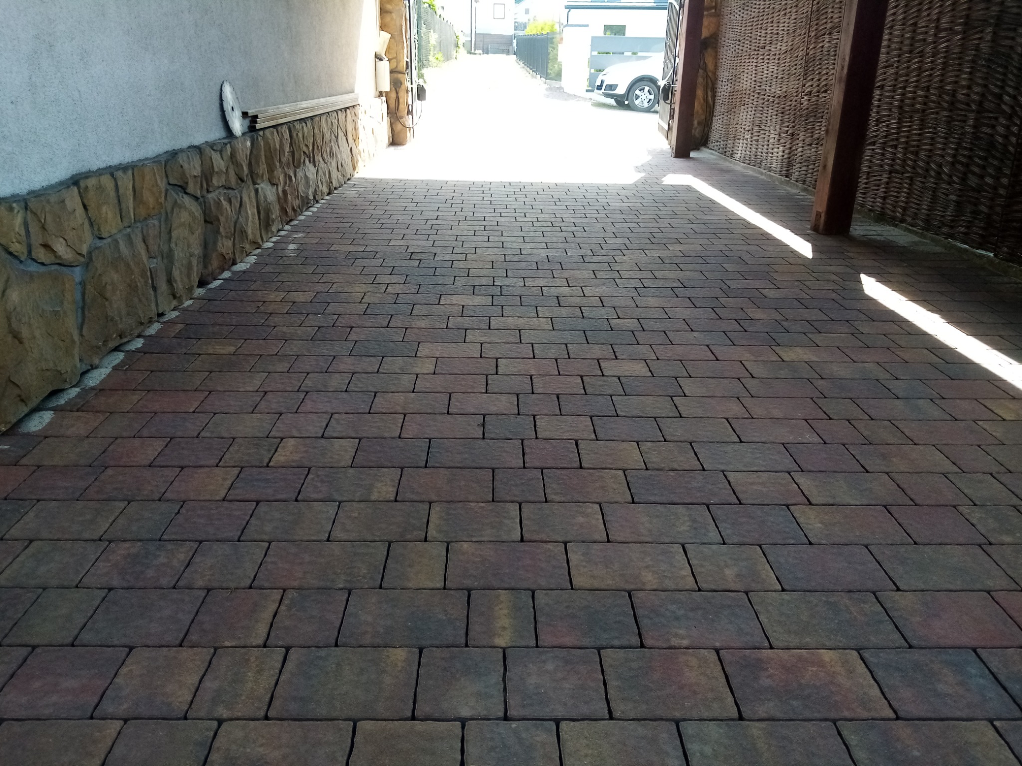 Ułożona kostka brukowa w odcieniach brązu i szarości prowadząca do jasnego wjazdu, z widocznym fragmentem ściany wykończonej kamieniem i elementami dekoracyjnymi.