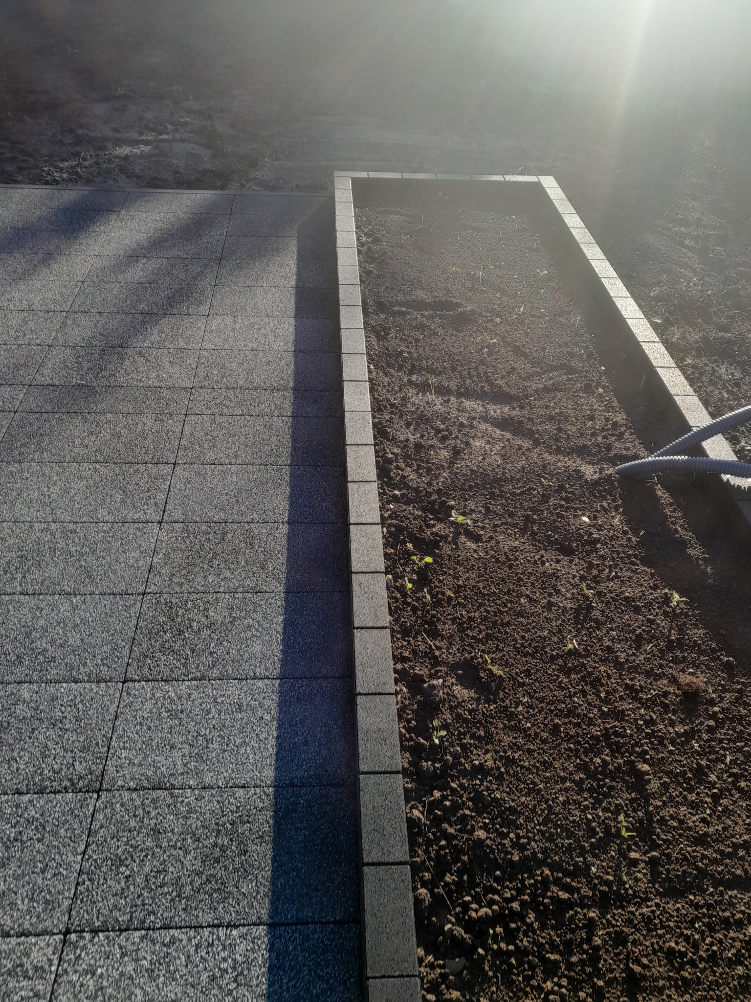 Ułożona szara kostka brukowa sąsiadująca z przygotowanym podwyższonym rabatem, obrzeże z tej samej kostki, w tle ziemia i wąż ogrodowy.