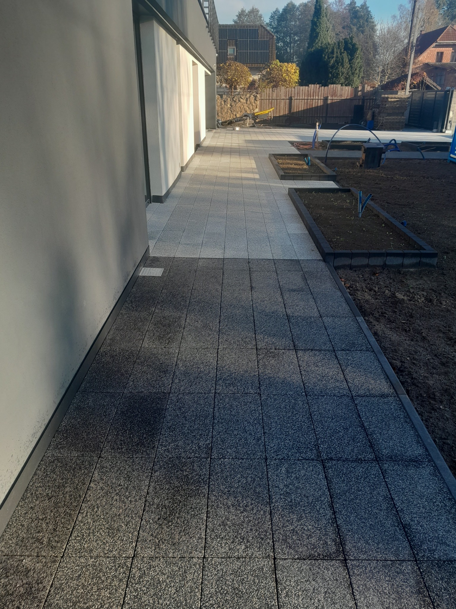 Ułożona dwukolorowa nawierzchnia z kostki brukowej przy nowoczesnym domu, z widocznymi rabatami kwiatowymi i elementami instalacji nawadniającej.