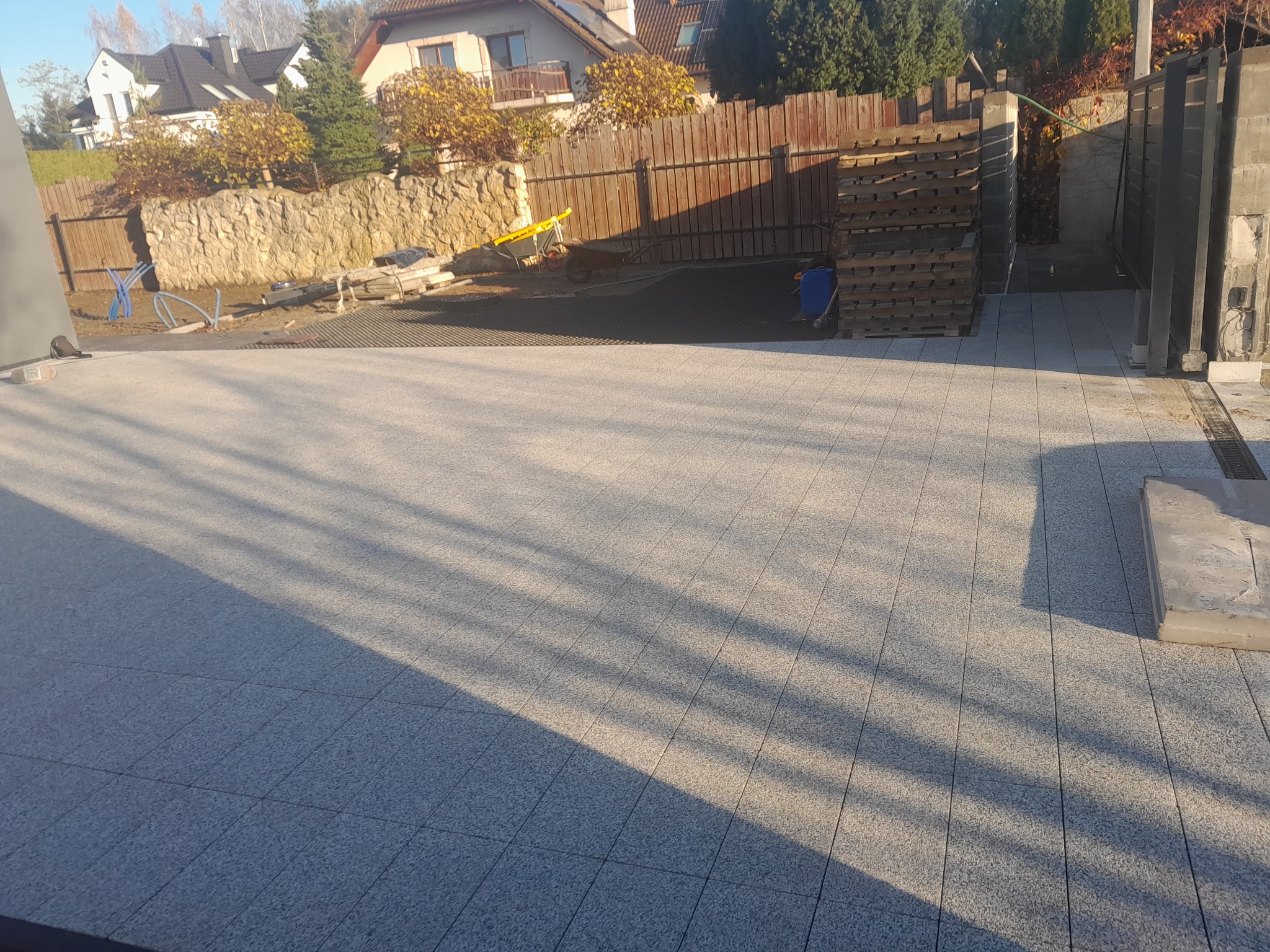 Ułożona jasnoszara nawierzchnia z kostki, widoczne palety, taczka i przygotowane podłoże pod dalsze prace, w tle ogrodzenie i dom.
