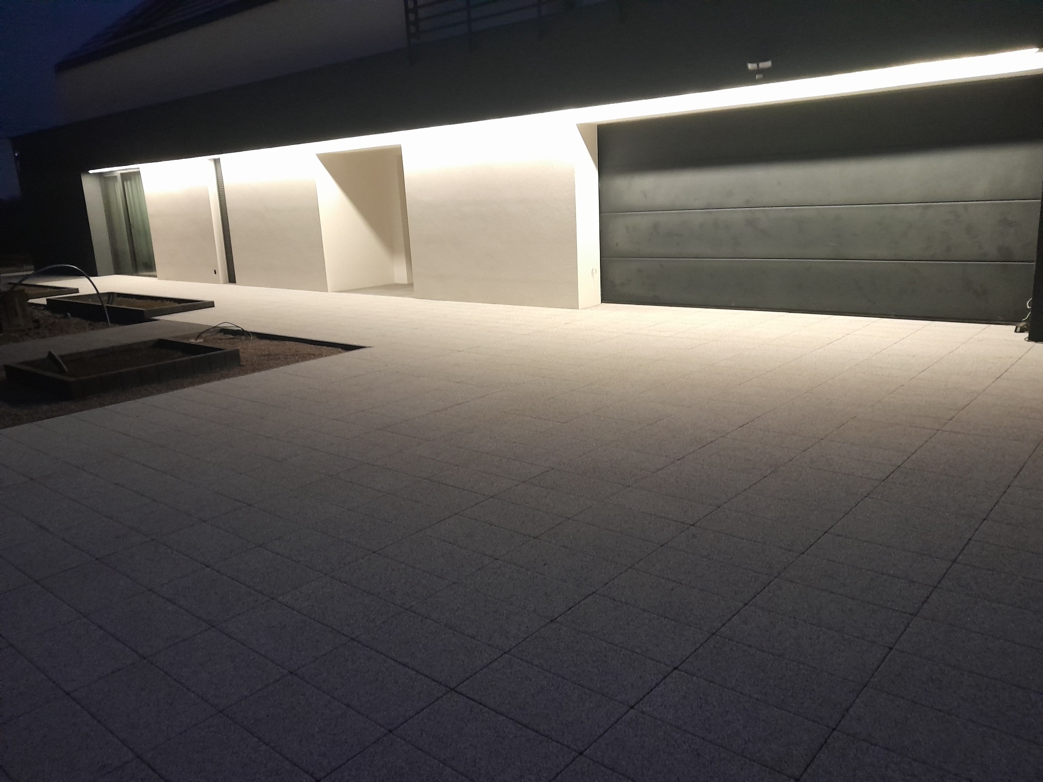 Nowoczesny podjazd z szarych kostek brukowych oświetlony przez zewnętrzne lampy, widok na bramę garażową i minimalistyczną architekturę domu.