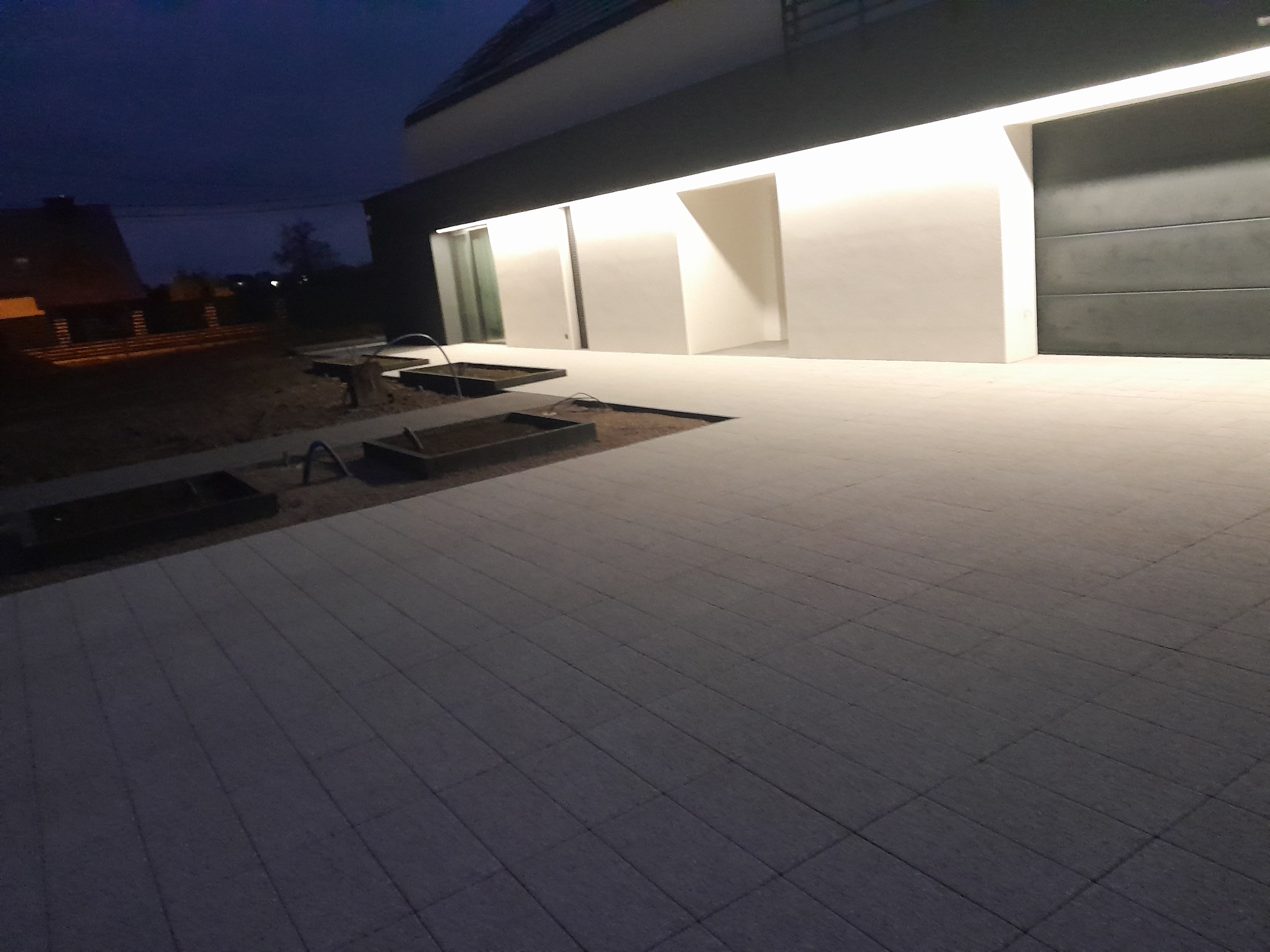 Nowoczesny podjazd z szarej kostki brukowej oświetlony nocą, z minimalistycznymi rabatami wzdłuż ścieżki i elewacją budynku z podświetleniem.