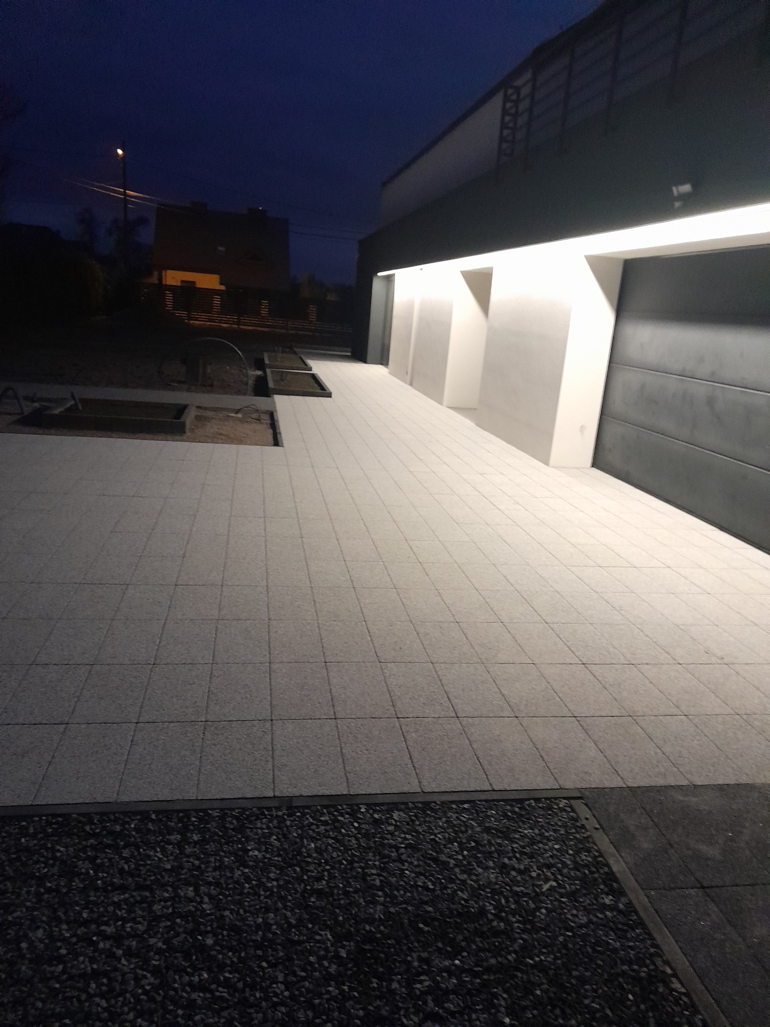 Nowoczesny podjazd z jasnoszarej kostki granitowej ułożonej przed garażem z podświetleniem, widoczne przygotowane rabaty ziemne i fragment dekoracyjnego obsypania z kamieni.