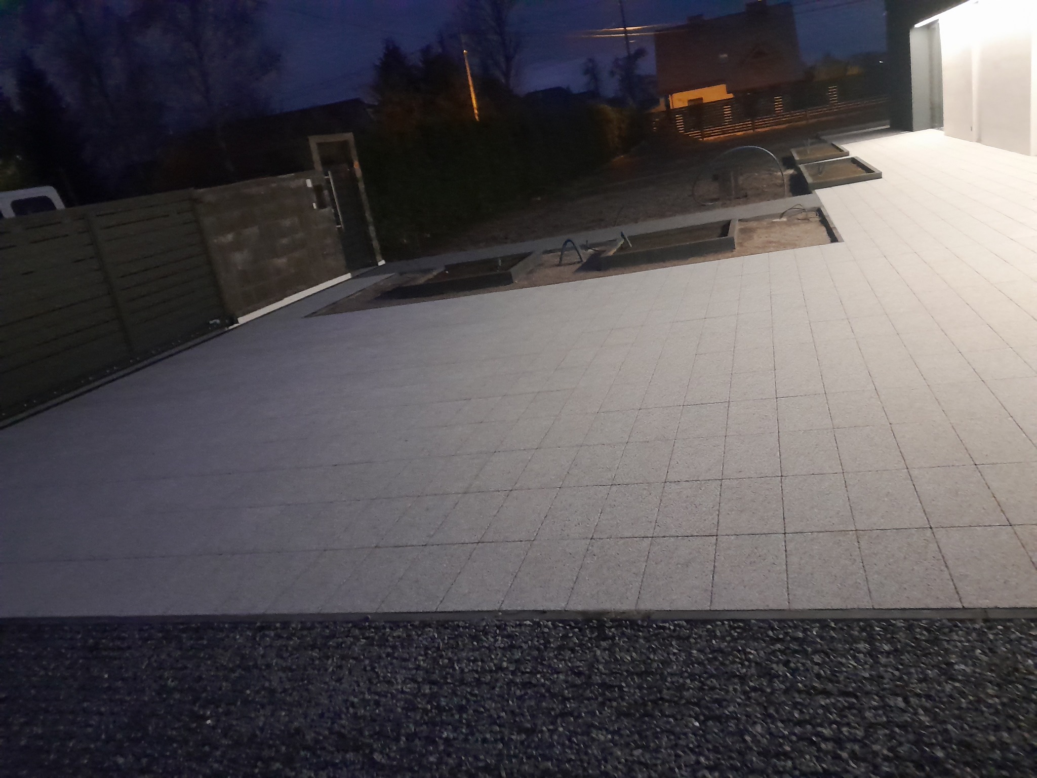 Nowa, jasnoszara nawierzchnia z kostki brukowej ułożona przed budynkiem w ciemnych barwach nocy, z widocznymi fragmentami gruntu przygotowanymi pod nasadzenia.