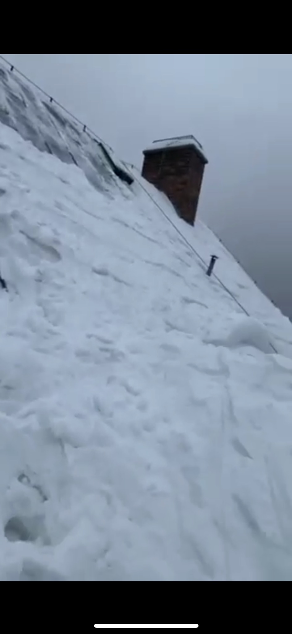 Zasypany śniegiem dach z ceglanym kominem w pochmurny dzień. Widoczne liny zabezpieczające.