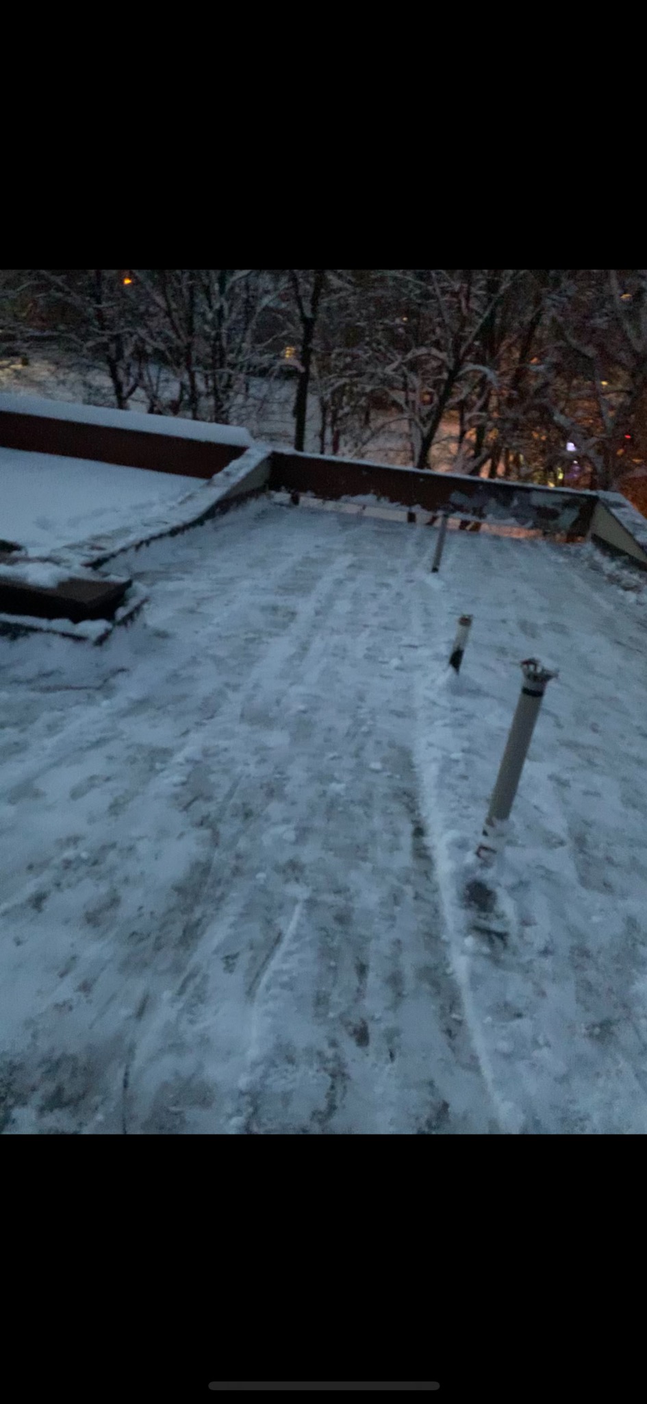 Śnieżny dach budynku w Lubieniu po odśnieżaniu, widoczne ślady odśnieżania i elementy konstrukcyjne dachu wystające spod śniegu, w tle drzewa i oświetlenie uliczne.