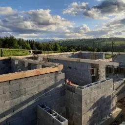 Wznoszone ściany fundamentowe z bloczków betonowych, z widocznym zbrojeniem i drewnianymi elementami szalunku, na tle malowniczego krajobrazu z górami i zachmurzonym niebem.