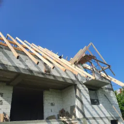 Surowy dom murowany z betonu komórkowego w trakcie budowy dachu, widoczne krokwie i rusztowanie, błękitne niebo w tle.