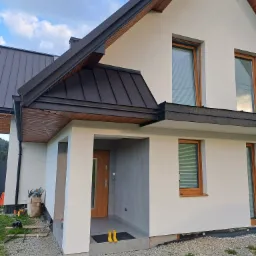Nowoczesny dom z białą elewacją, ciemnym dachem z blachy na rąbek i drewnianymi oknami, żółte kalosze przy wejściu, teren wokół domu częściowo wyłożony kamieniem.