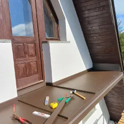 Blacha na dachu lukarny, narzędzia dekarskie, widok z góry na fragment dachu z oknem i drewnianą boazerią, etap prac dekarskich.