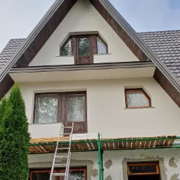 Dom jednorodzinny w trakcie prac elewacyjnych, widoczne rusztowanie, drabina oparta o ścianę, częściowo ocieplona ściana zewnętrzna z widocznymi fragmentami styropianu, okna z drewnianą stolarką...