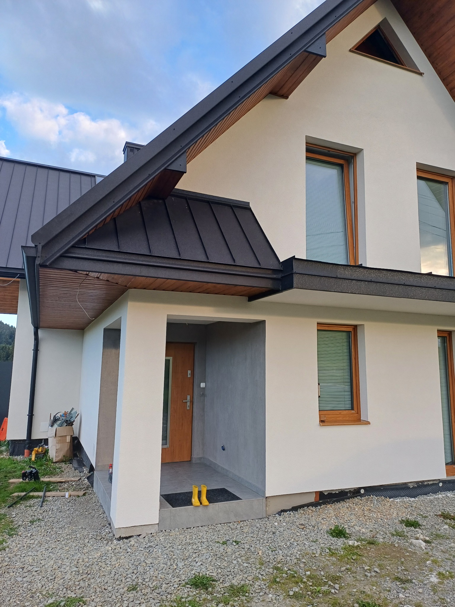 Nowoczesny dom z białą elewacją, ciemnym dachem z blachy na rąbek i drewnianymi oknami, żółte kalosze przy wejściu, teren wokół domu częściowo wyłożony kamieniem.