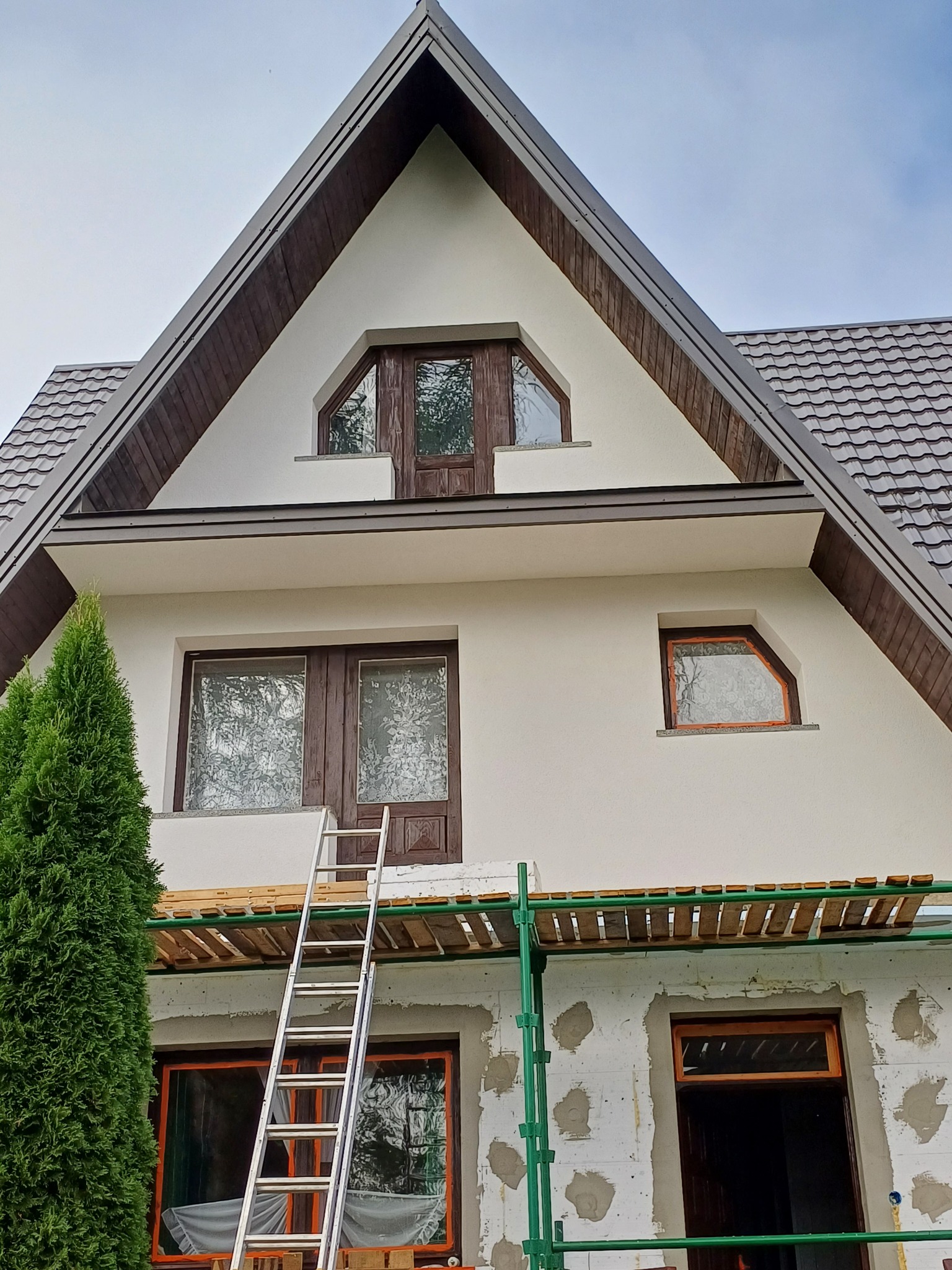 Dom jednorodzinny w trakcie prac elewacyjnych, widoczne rusztowanie, drabina oparta o ścianę, częściowo ocieplona ściana zewnętrzna z widocznymi fragmentami styropianu, okna z drewnianą stolarką...