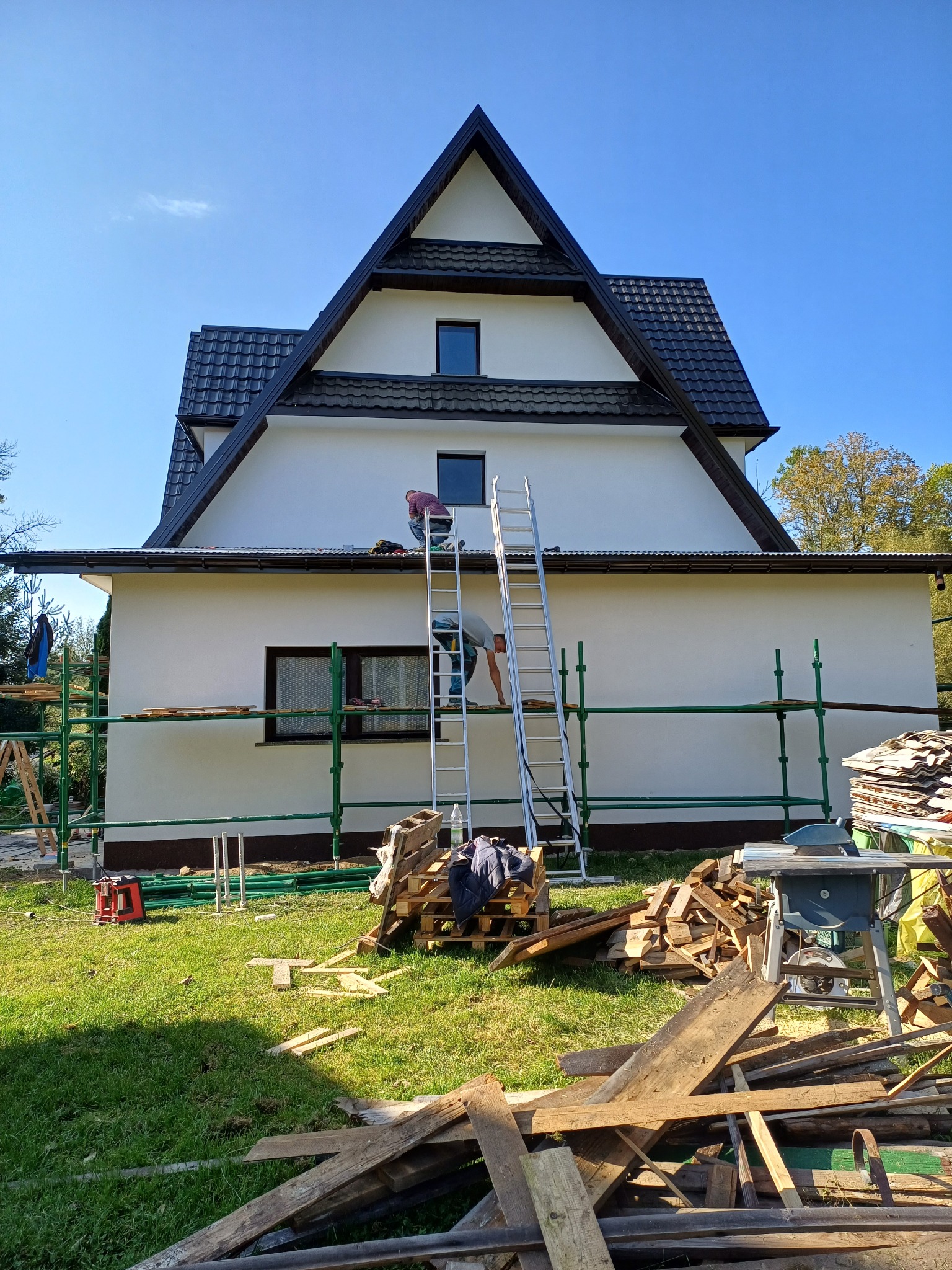 Dwóch mężczyzn pracuje na dachu domu z białymi ścianami, używając rusztowania i drabiny; na trawniku leżą deski i narzędzia. Widoczny fragment dachu z ciemną dachówką.