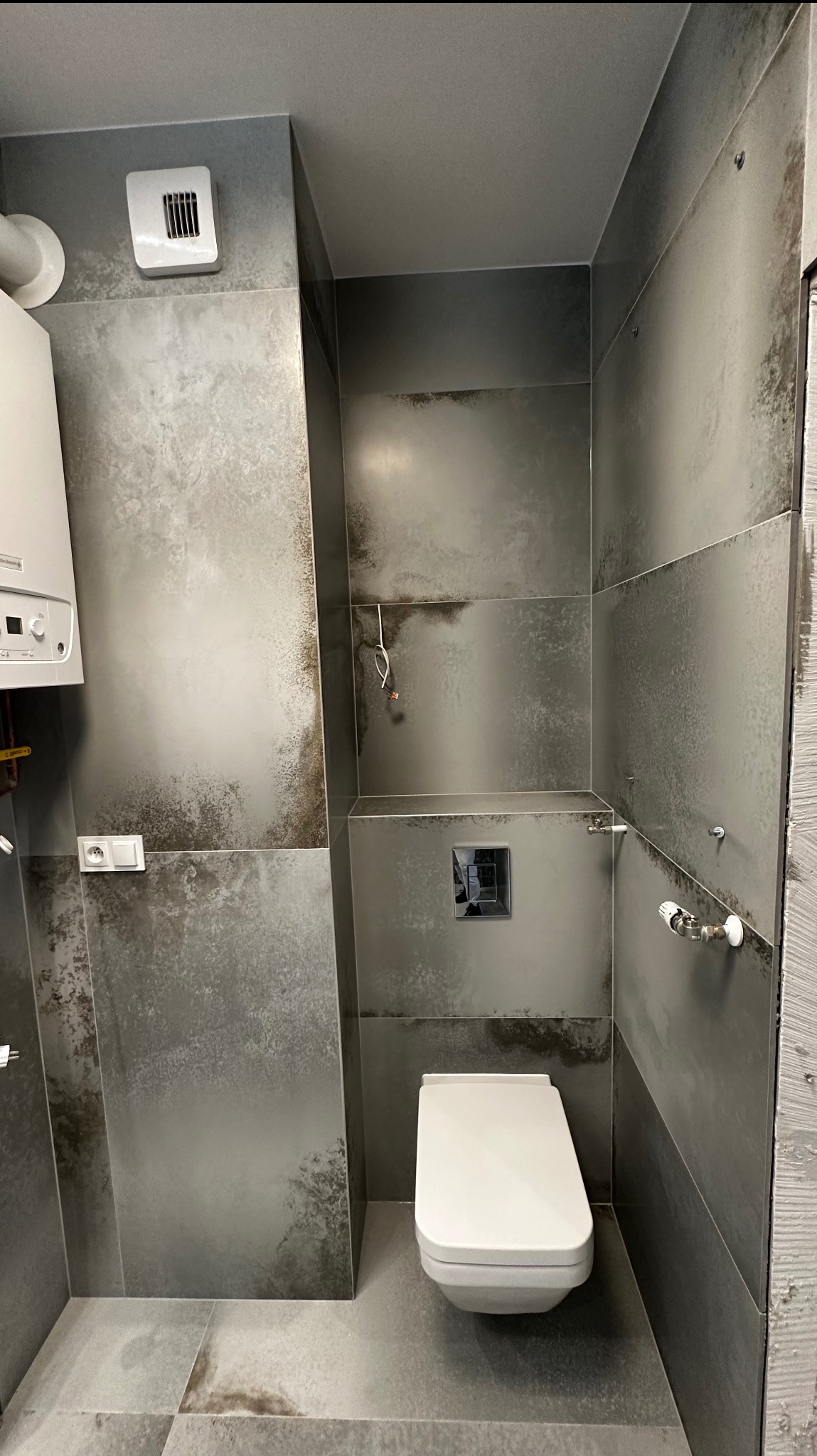 Nowoczesna łazienka w szarościach z podtynkową toaletą, płytki imitujące metal, wiszący bojler, widoczne przewody elektryczne i rury hydrauliczne przed zabudową.