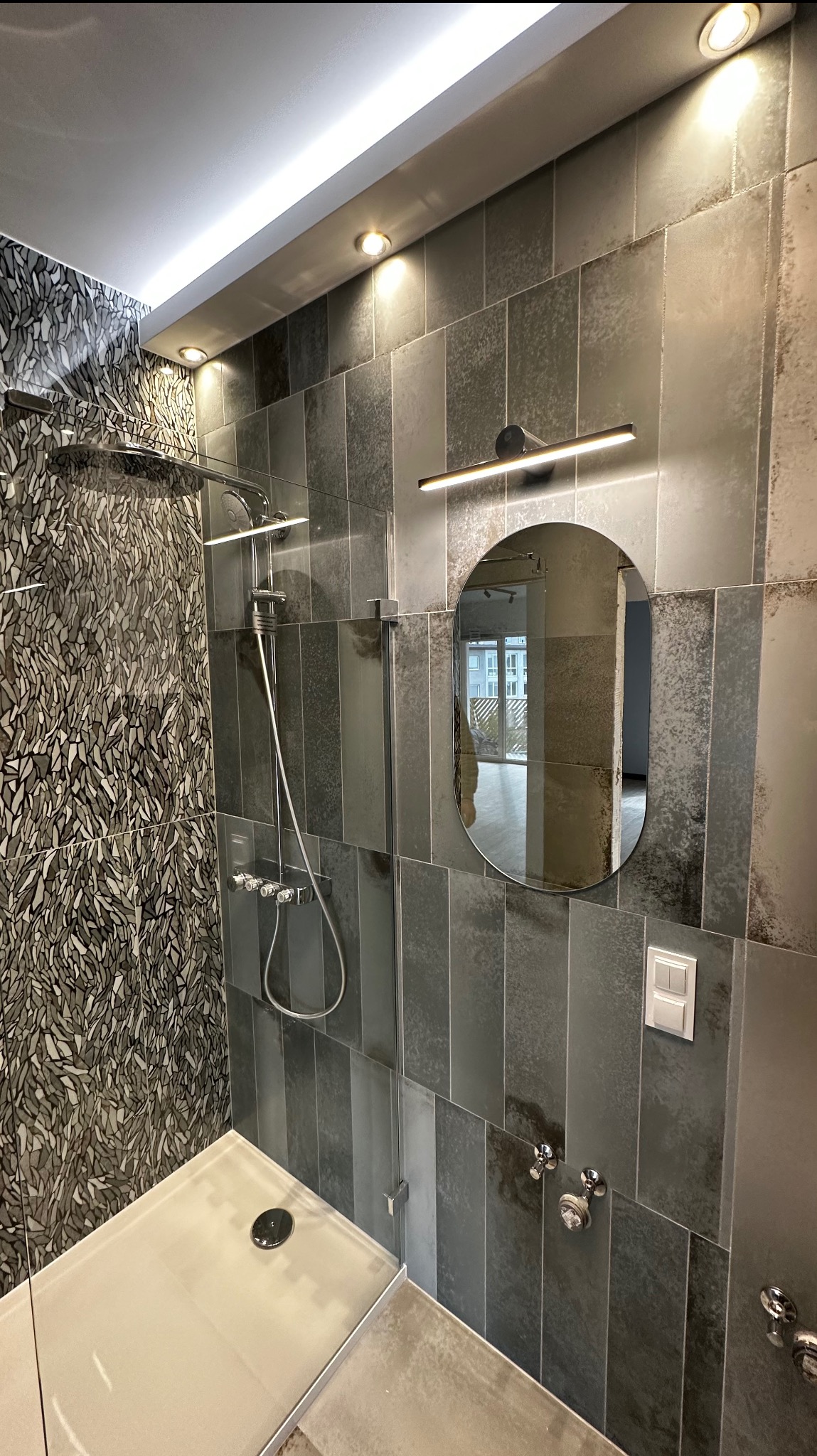Nowoczesna kabina prysznicowa z deszczownicą i szklaną ścianką, obok owalne lustro z oświetleniem LED na ścianie z szarych płytek w pionowym układzie, mozaika na ścianie prysznica