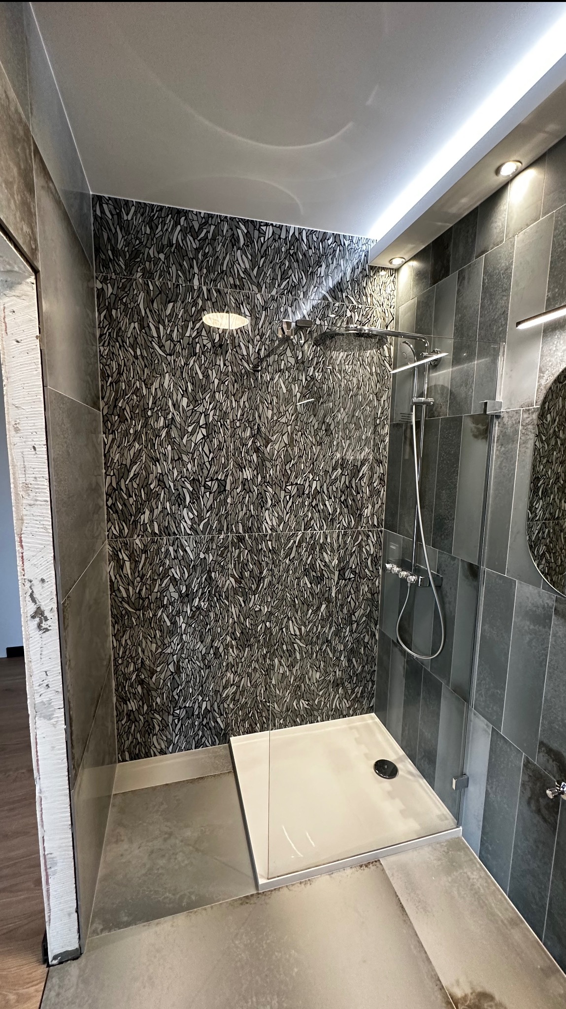 Nowoczesna kabina prysznicowa z deszczownicą, wykończona płytkami w odcieniach szarości, z akcentem wzoru liści na jednej ze ścian, oraz subtelnym oświetleniem LED.