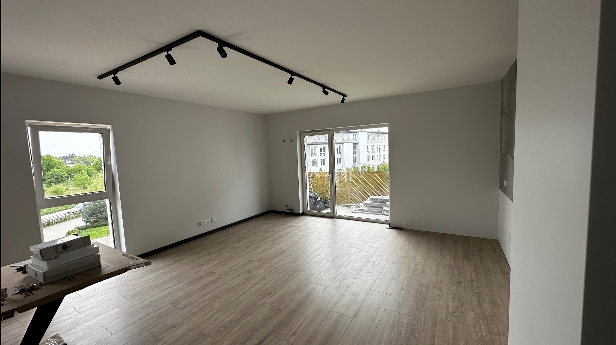 Wnętrze pustego pokoju po remoncie z panelami podłogowymi w odcieniach beżu, czarnym oświetleniem szynowym na suficie i oknem z widokiem na zieleń. Na pierwszym planie kartony ze sprzętem.