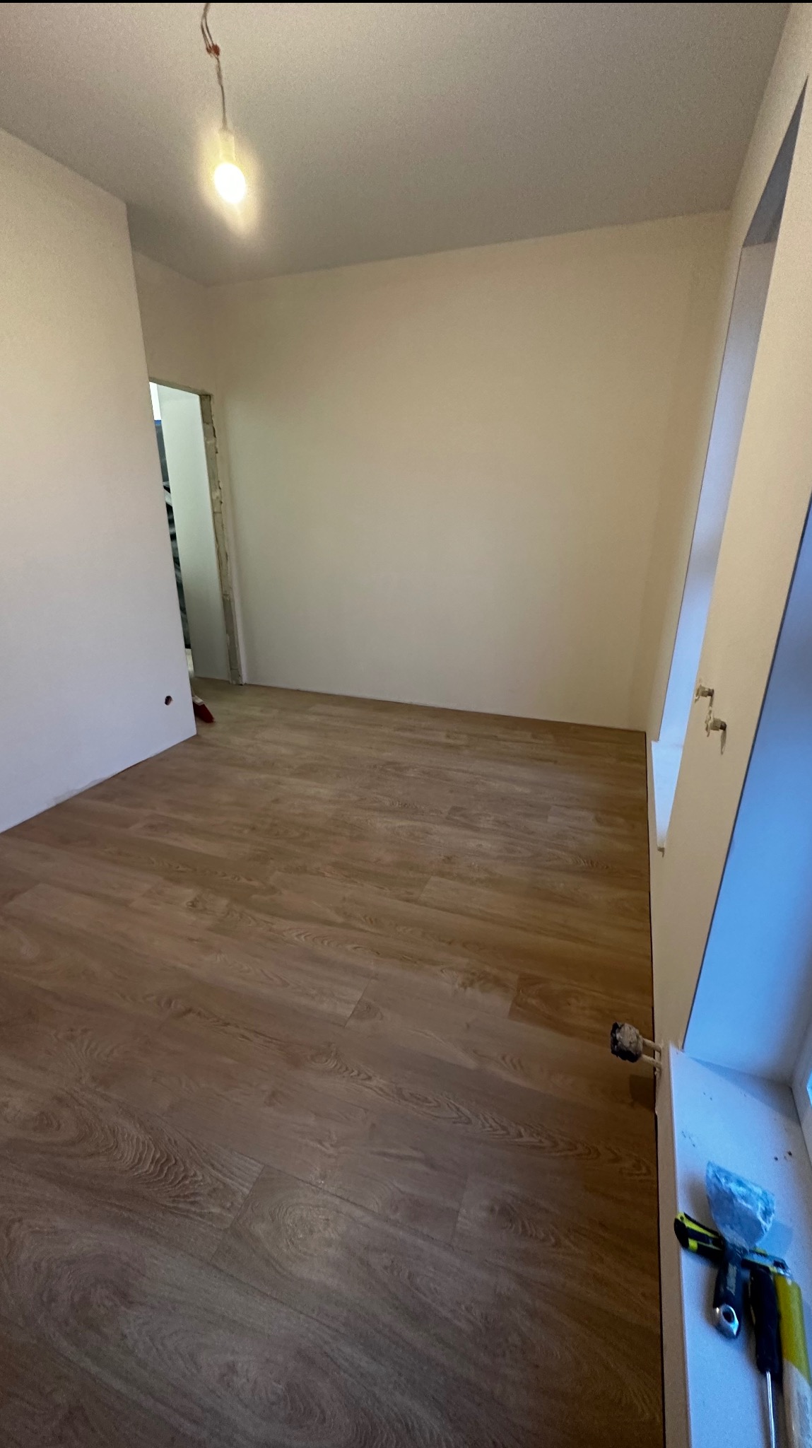 Pusta przestrzeń z nowo ułożonymi panelami podłogowymi o wzorze drewna, surowe ściany pomalowane na biało, widoczna instalacja elektryczna pod sufitem, narzędzia na parapecie okiennym.