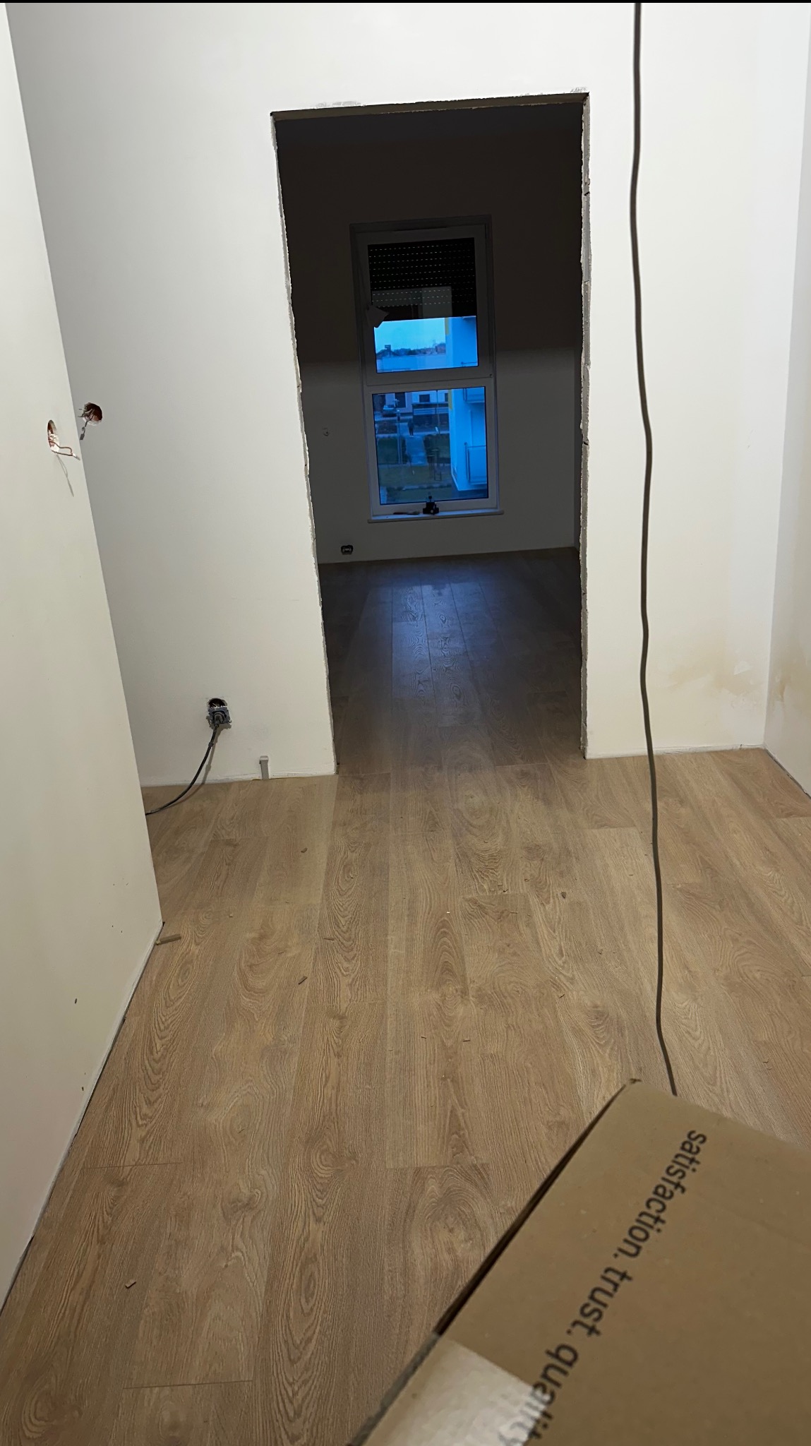 Wnętrze w trakcie wykańczania: świeżo położone panele podłogowe w odcieniach drewna, białe ściany z widocznymi punktami elektrycznymi, widok na okno z roletami i budynki na zewnątrz, fragment...