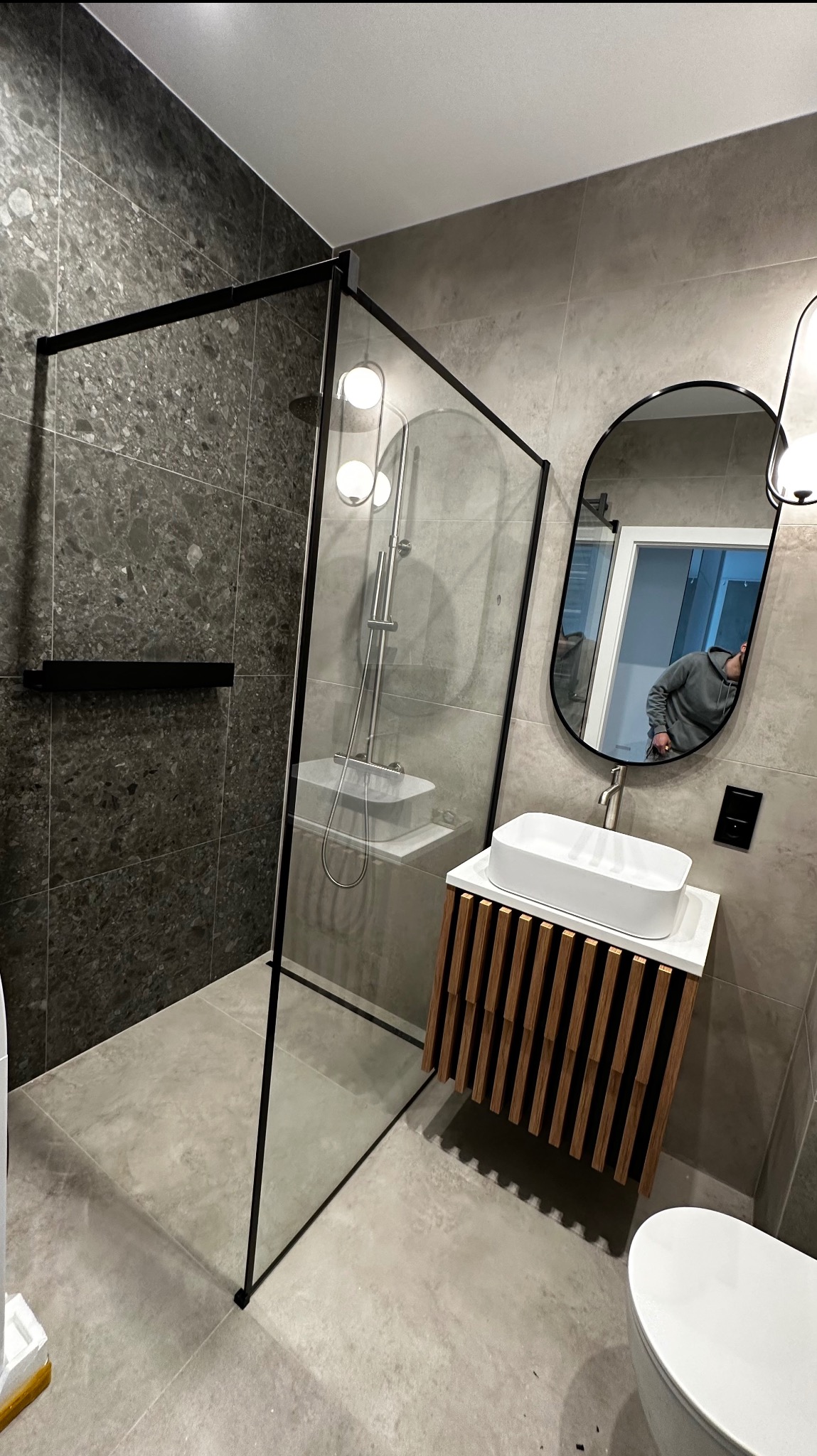 Nowoczesna łazienka z kabiną prysznicową typu walk-in z czarnymi ramami, umywalką nablatową na szafce z drewnianych listewek i owalnym lustrem z odbiciem fotografa.