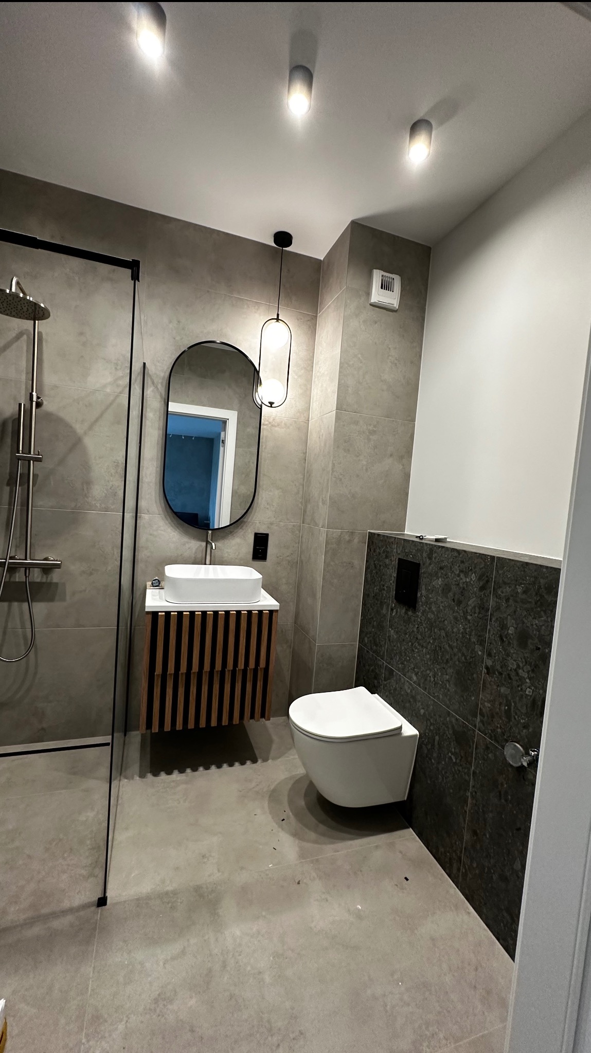 Nowoczesna łazienka z szarymi płytkami, owalnym lustrem z czarną ramą, wiszącą toaletą i umywalką na szafce z drewnianymi lamelami, oświetlona wiszącą lampą z kulistymi kloszami.