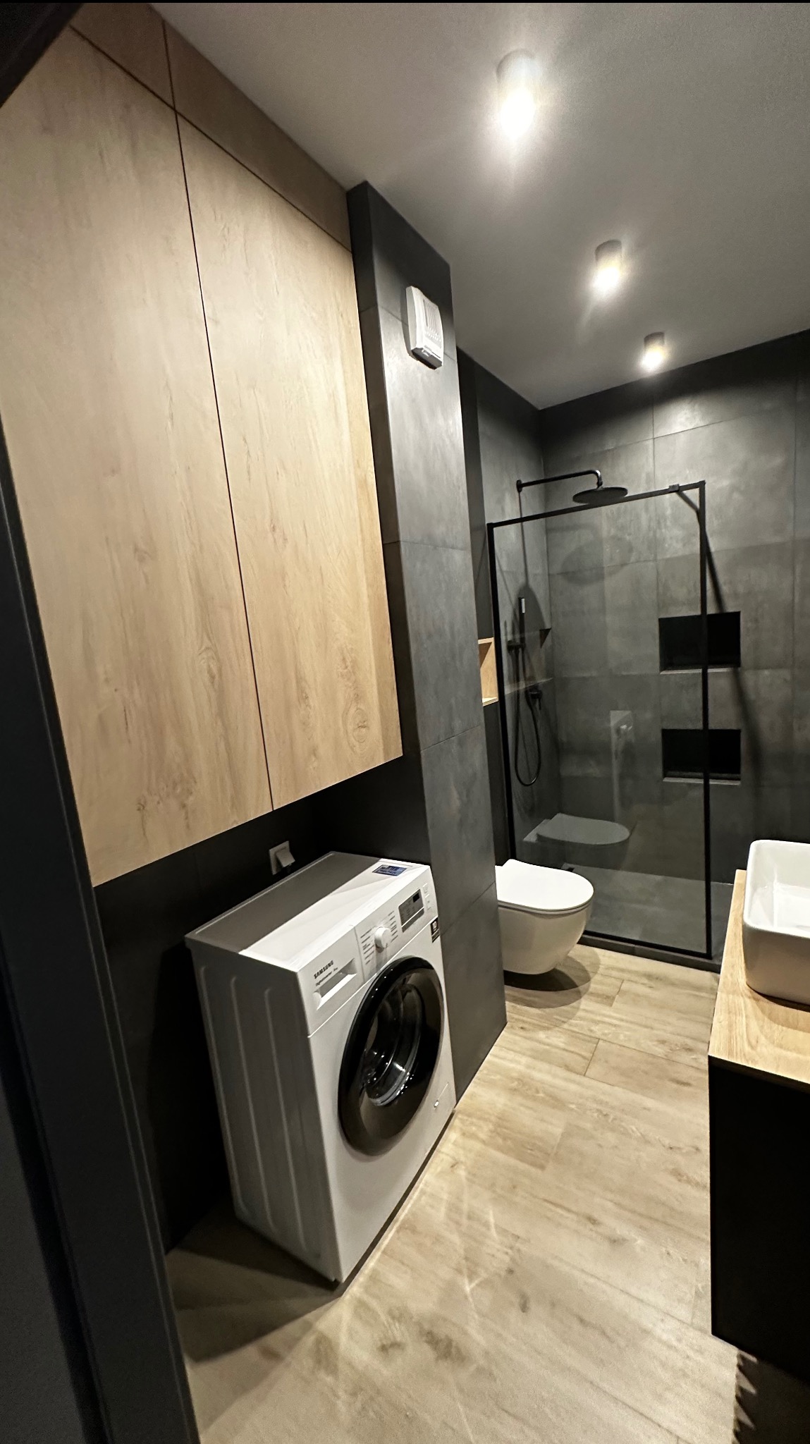 Nowoczesna łazienka z kabiną prysznicową typu walk-in, wiszącą toaletą, pralką Samsung i drewnianymi szafkami pod sufitem, wykończona szarymi płytkami i panelami podłogowymi imitującymi drewno.