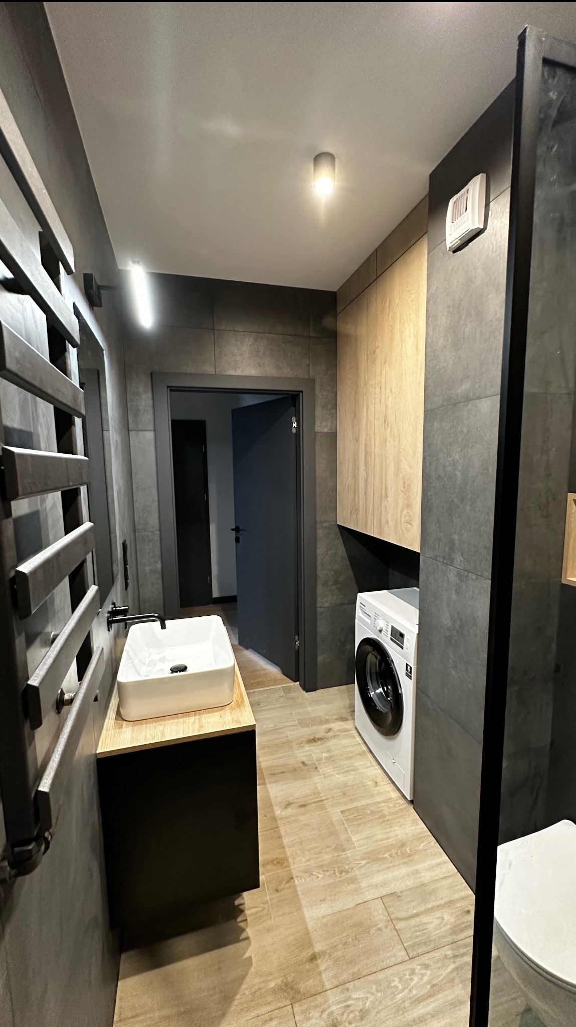 Nowoczesna łazienka z ciemnymi płytkami, drewnopodobną podłogą, białą umywalką nablatową na czarnej szafce, grzejnikiem drabinkowym, pralką i toaletą.