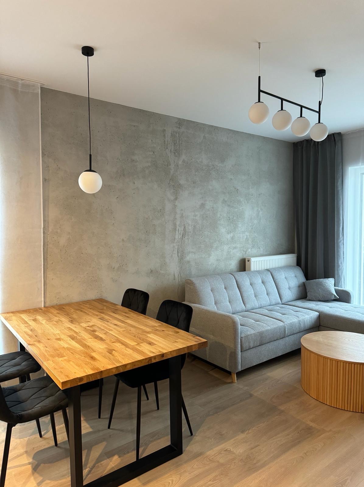 Nowoczesny salon z drewnianym stołem, czarnymi krzesłami, szarą sofą narożną i betonową ścianą akcentową, oświetlony kulistymi lampami wiszącymi.