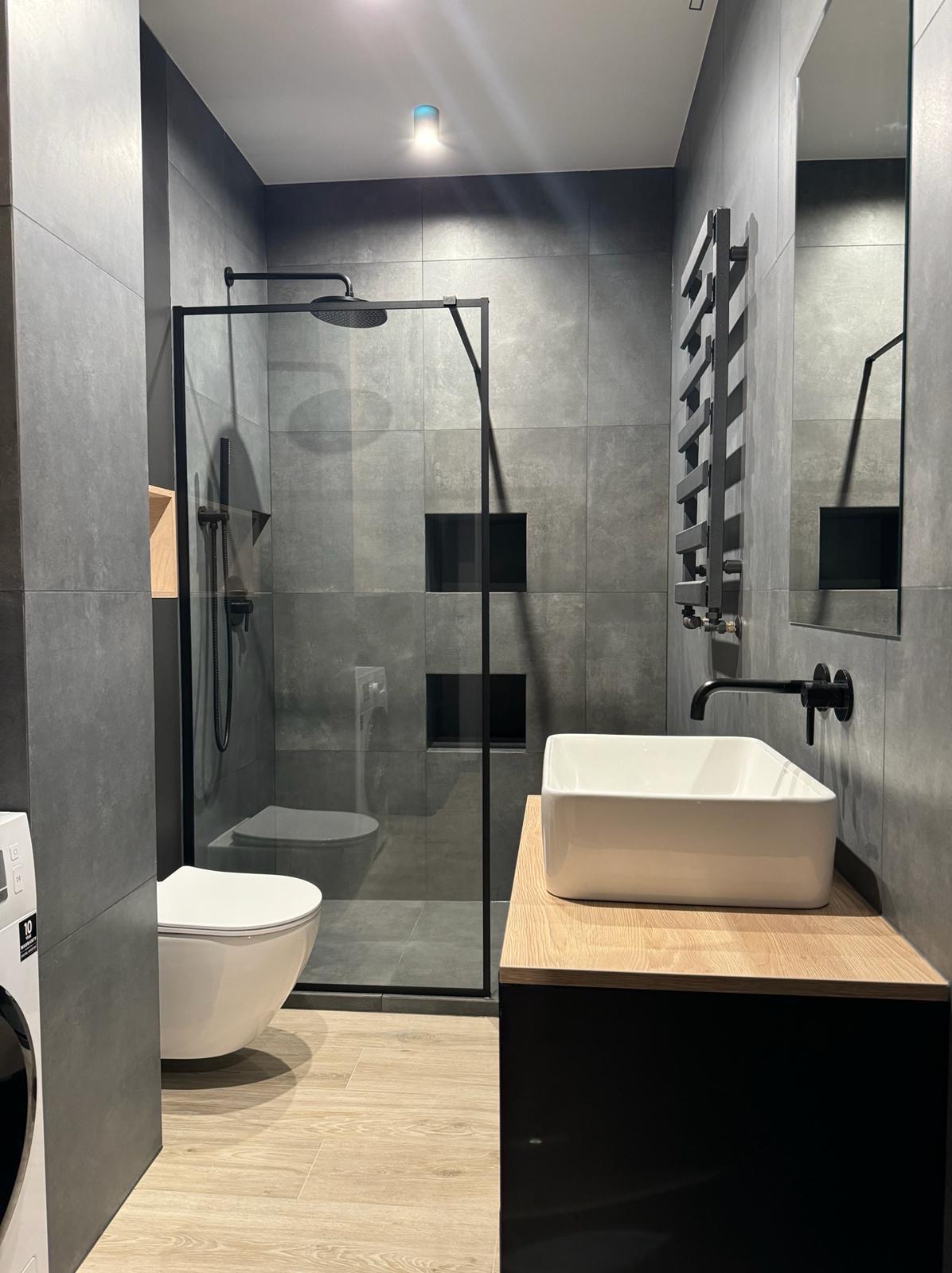 Nowoczesna łazienka z szarymi płytkami, kabiną prysznicową typu walk-in z czarnymi ramami, wiszącą toaletą, białym umywalką nablatową na drewnianym blacie i czarną armaturą.