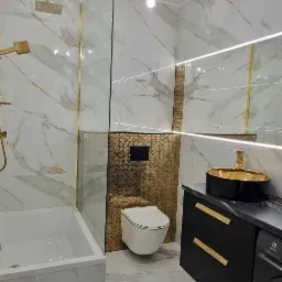 Elegancka łazienka z marmurowymi płytkami, złotymi akcentami, czarną szafką z umywalką i podwieszaną toaletą na tle złotej mozaiki.