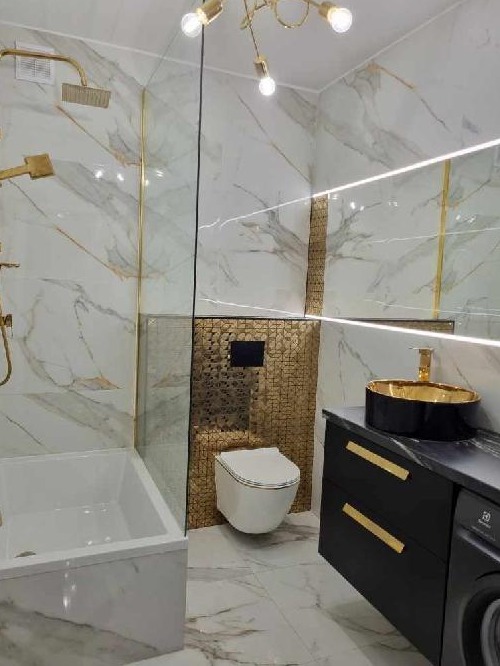Elegancka łazienka z marmurowymi płytkami, złotymi akcentami, czarną szafką z umywalką i podwieszaną toaletą na tle złotej mozaiki.