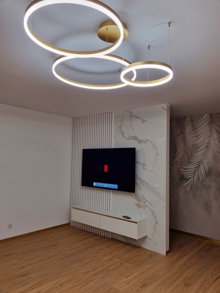 Nowoczesny salon z podświetlanymi okrągłymi lampami sufitowymi, telewizorem na ścianie z dekoracyjnymi panelami pionowymi i marmurową płytą, oraz niską białą szafką RTV.