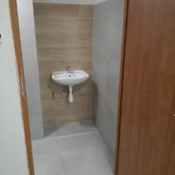 Niewielka łazienka po remoncie: beżowe płytki imitujące drewno za białą umywalką, jasnoszare płytki na ścianach bocznych i podłodze, widoczna część drewnianych drzwi.