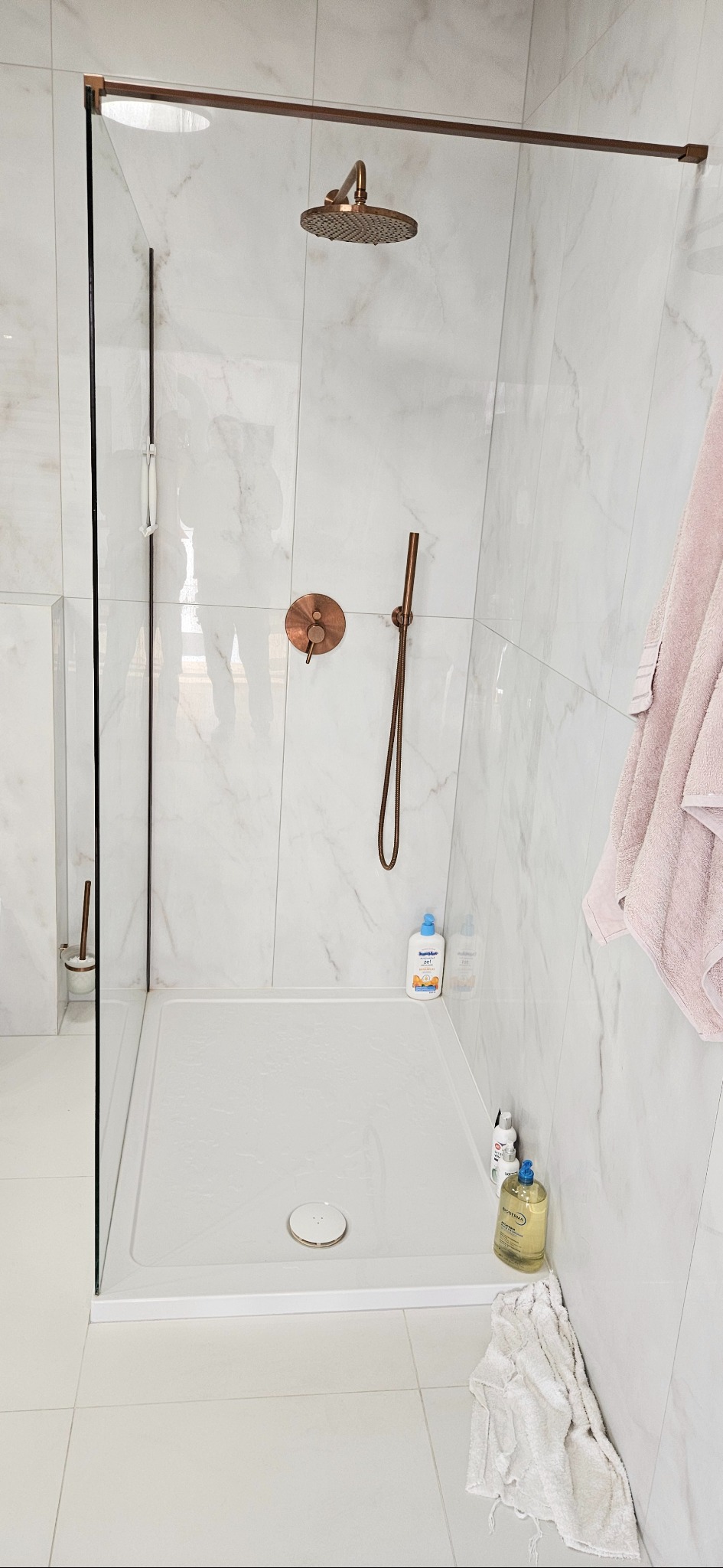 Nowoczesna kabina prysznicowa z marmurowymi płytkami, armaturą w kolorze miedzi i szklanymi drzwiami. Widoczne kosmetyki i ręcznik. Minimalistyczny design.