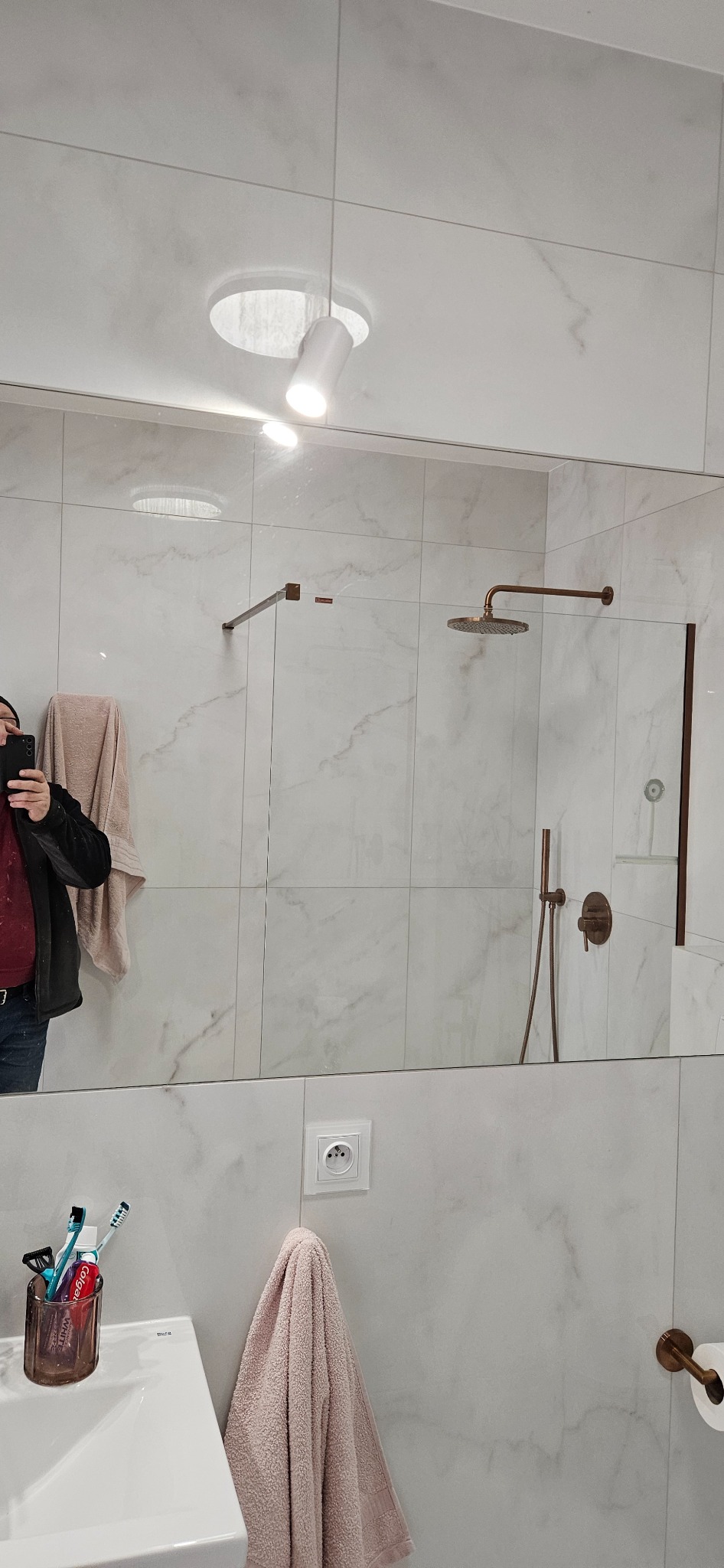 Nowoczesna łazienka z marmurowymi płytkami, armaturą w kolorze miedzi i oświetleniem punktowym. Widoczne odbicie osoby robiącej zdjęcie w lustrze.