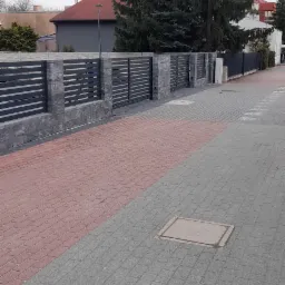 Nowoczesne ogrodzenie z poziomych, szarych paneli i kamiennych słupków, widoczne z chodnika wyłożonego kostką w dwóch kolorach, z drzewami iglastymi w tle.