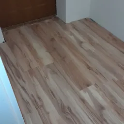 Ułożona podłoga laminowana w odcieniach beżu i brązu, imitująca drewno, widoczna przy drzwiach i białej ścianie w rogu pomieszczenia.