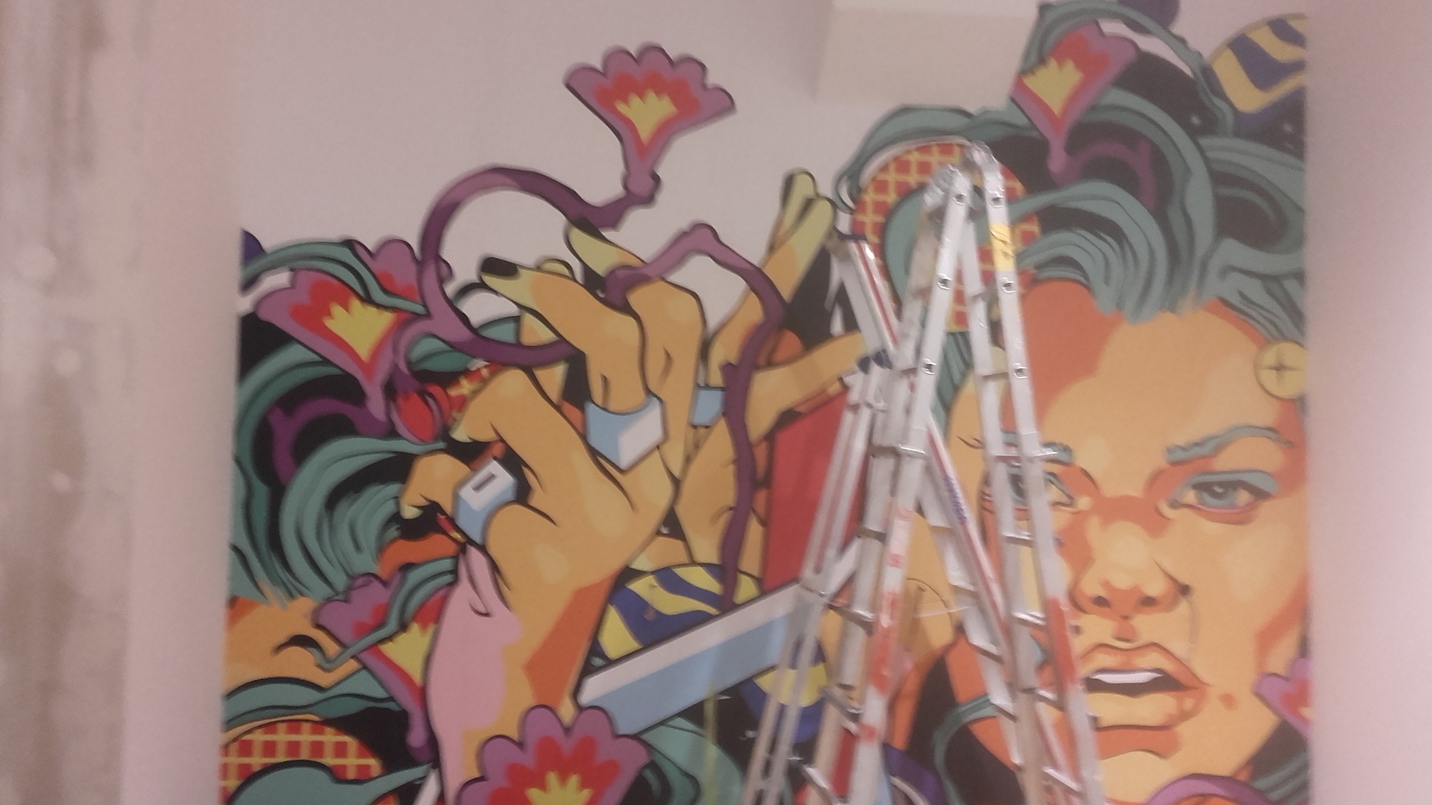 Kolorowy mural z motywami kwiatowymi i portretem kobiety, częściowo zasłonięty przez aluminiową drabinę rozstawną podczas prac wykończeniowych na ścianie.