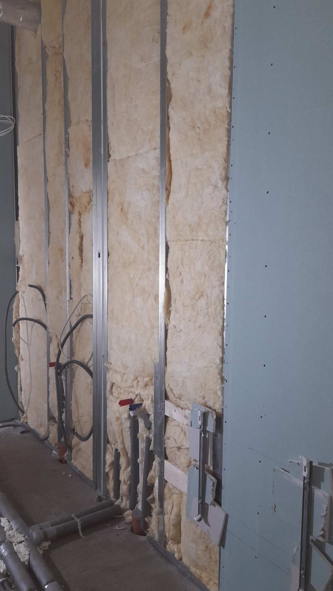 Ściana w trakcie izolacji termicznej z widoczną wełną mineralną między metalowymi profilami, instalacją hydrauliczną i częściowo zamontowaną płytą gipsowo-kartonową.