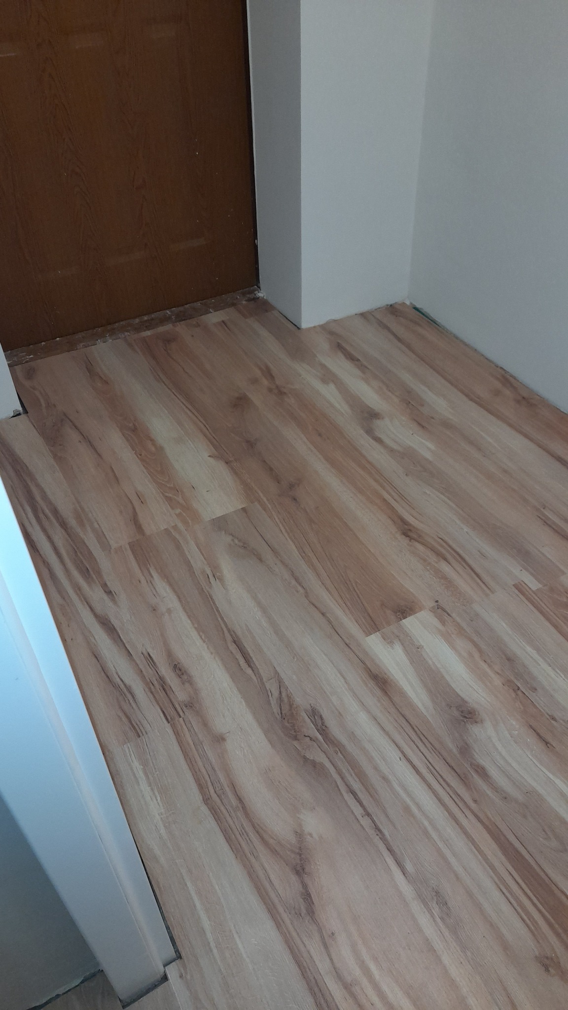 Ułożona podłoga laminowana w odcieniach beżu i brązu, imitująca drewno, widoczna przy drzwiach i białej ścianie w rogu pomieszczenia.