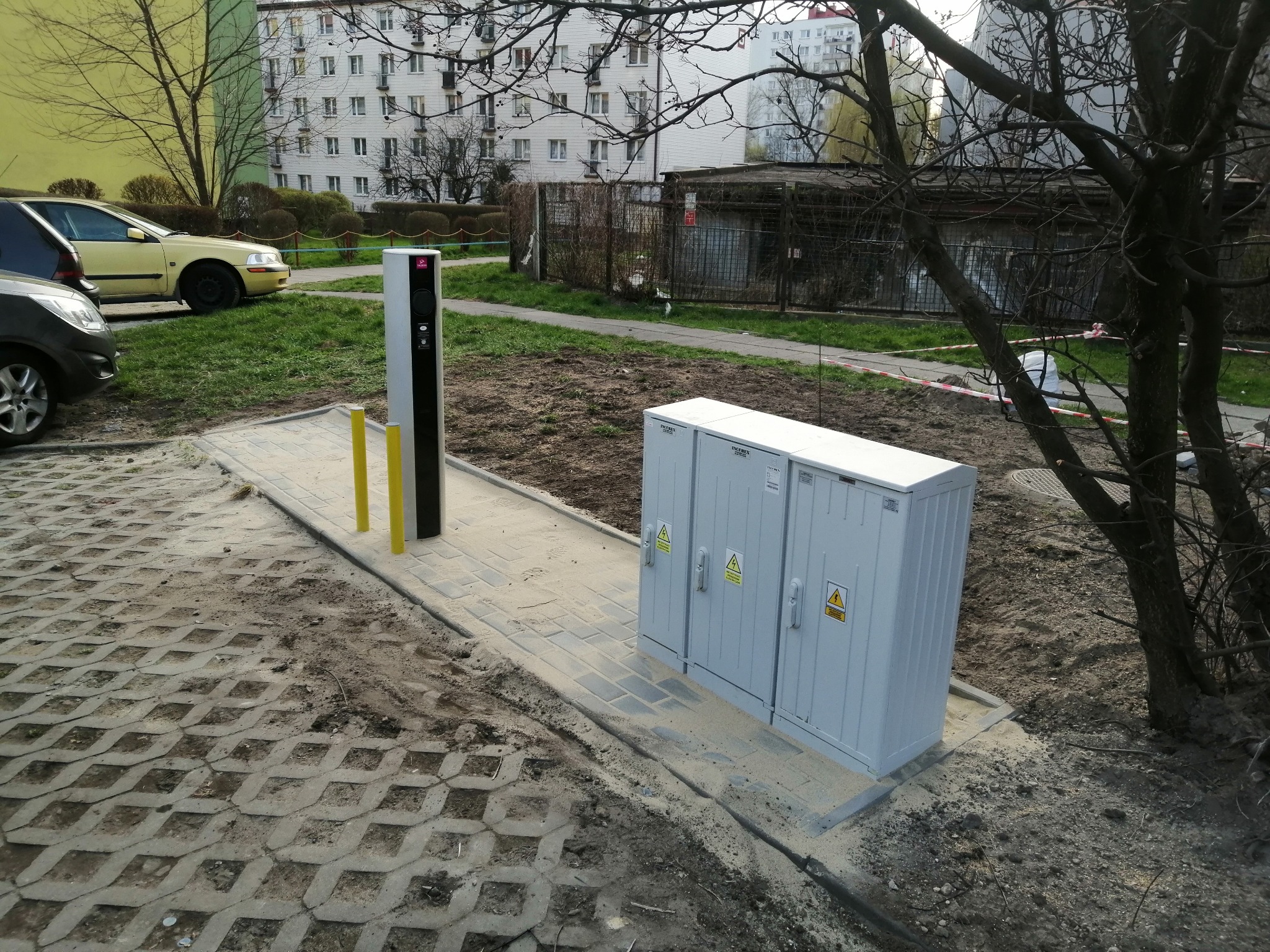 Nowo zainstalowana rozdzielnica elektryczna i słup ładowania pojazdów elektrycznych w Mysłowicach, na tle bloku mieszkalnego i drzew bez liści.