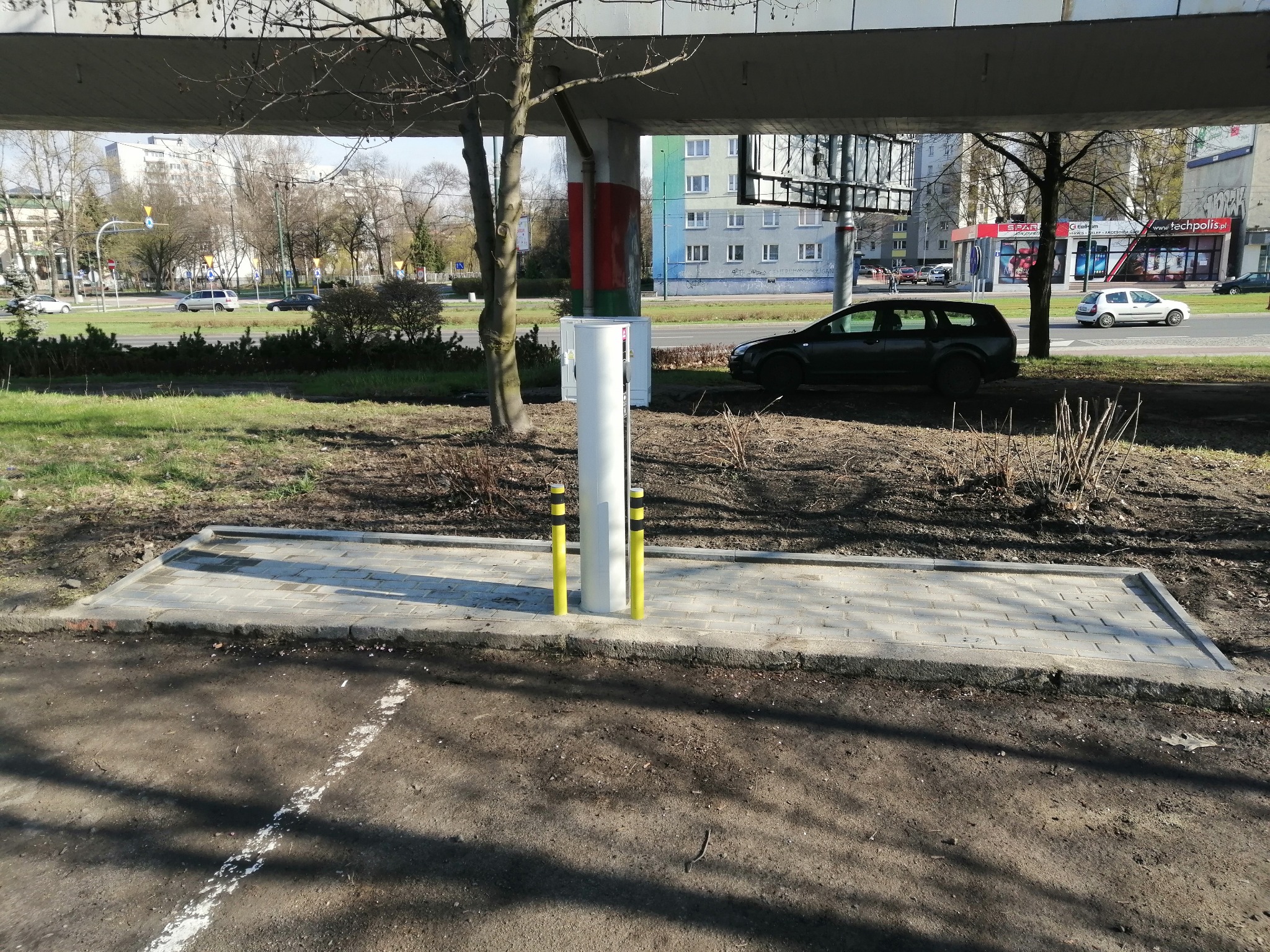 Nowa stacja ładowania pojazdów elektrycznych z dwoma słupkami ochronnymi, umieszczona na chodniku z kostki brukowej pod wiaduktem, w tle ulica i budynki.