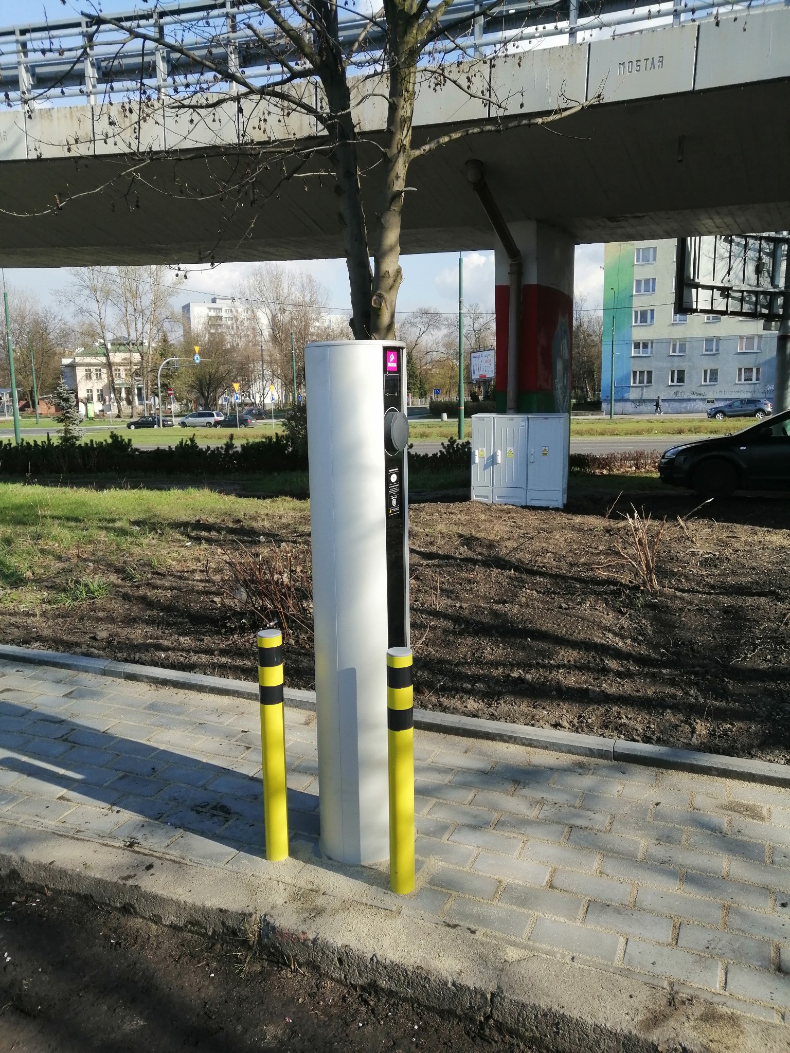 Biała stacja ładowania pojazdów elektrycznych z dwoma żółto-czarnymi słupkami zabezpieczającymi, umieszczona na chodniku w tle widoczny most i budynki.