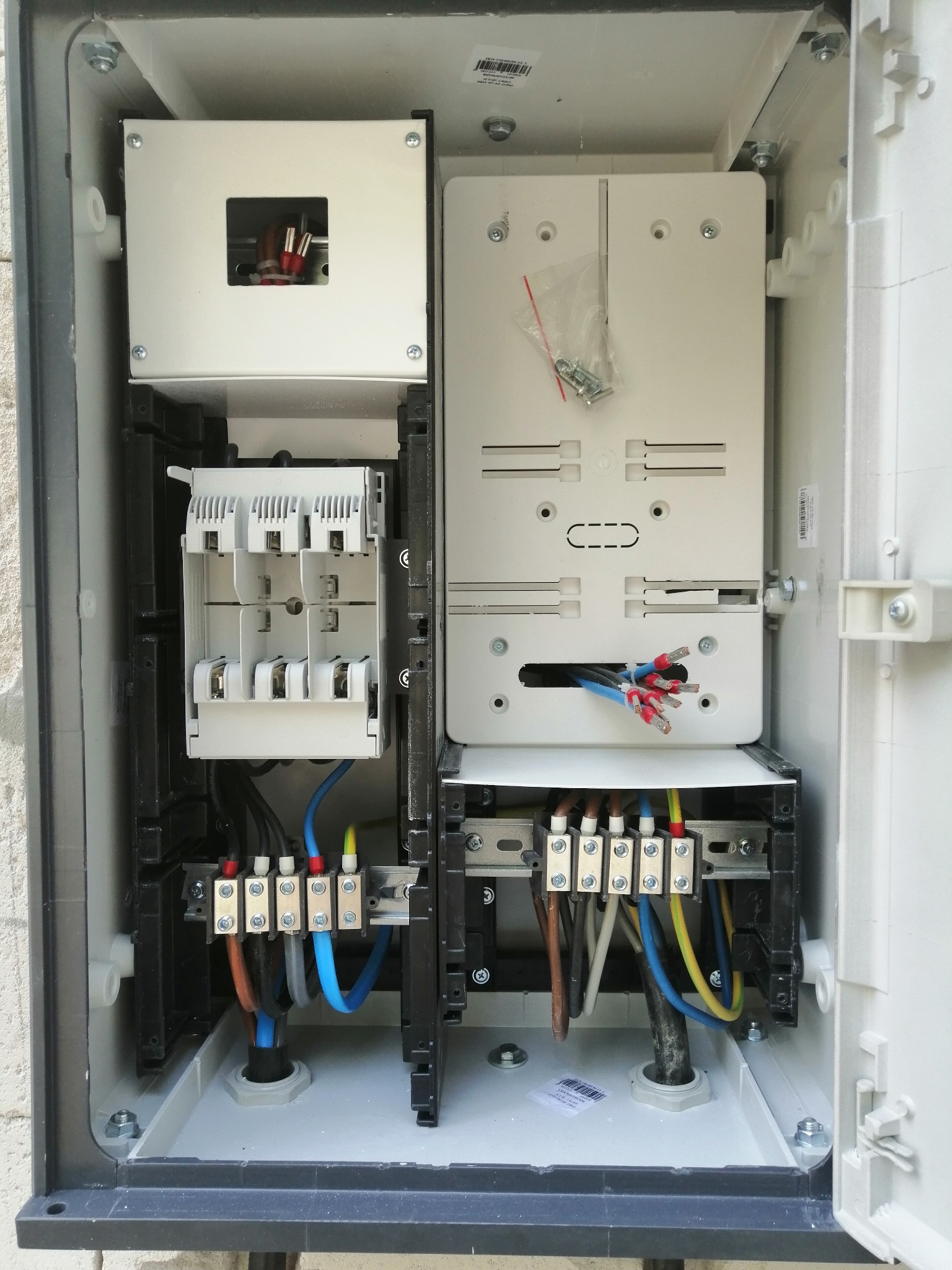 Otwarta skrzynka elektryczna z widocznymi bezpiecznikami, przewodami i listwami zaciskowymi, zainstalowana na szarej, chropowatej ścianie.