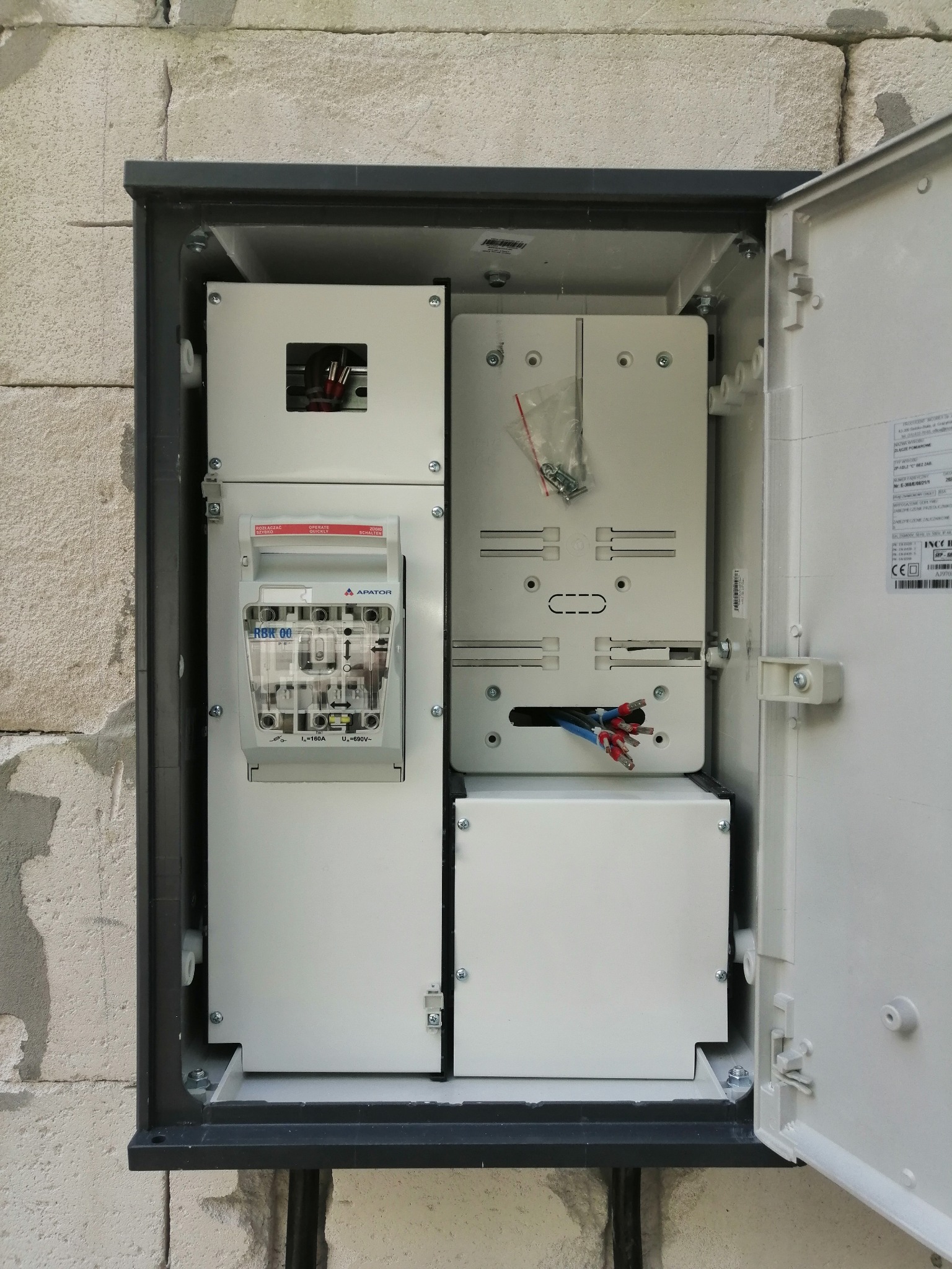 Otwarta skrzynka elektryczna zamontowana na ścianie z bloczków betonowych, widoczne elementy instalacji: wyłącznik APATOR RBK 00, przewody z końcówkami tulejkowymi oraz woreczek ze śrubami.