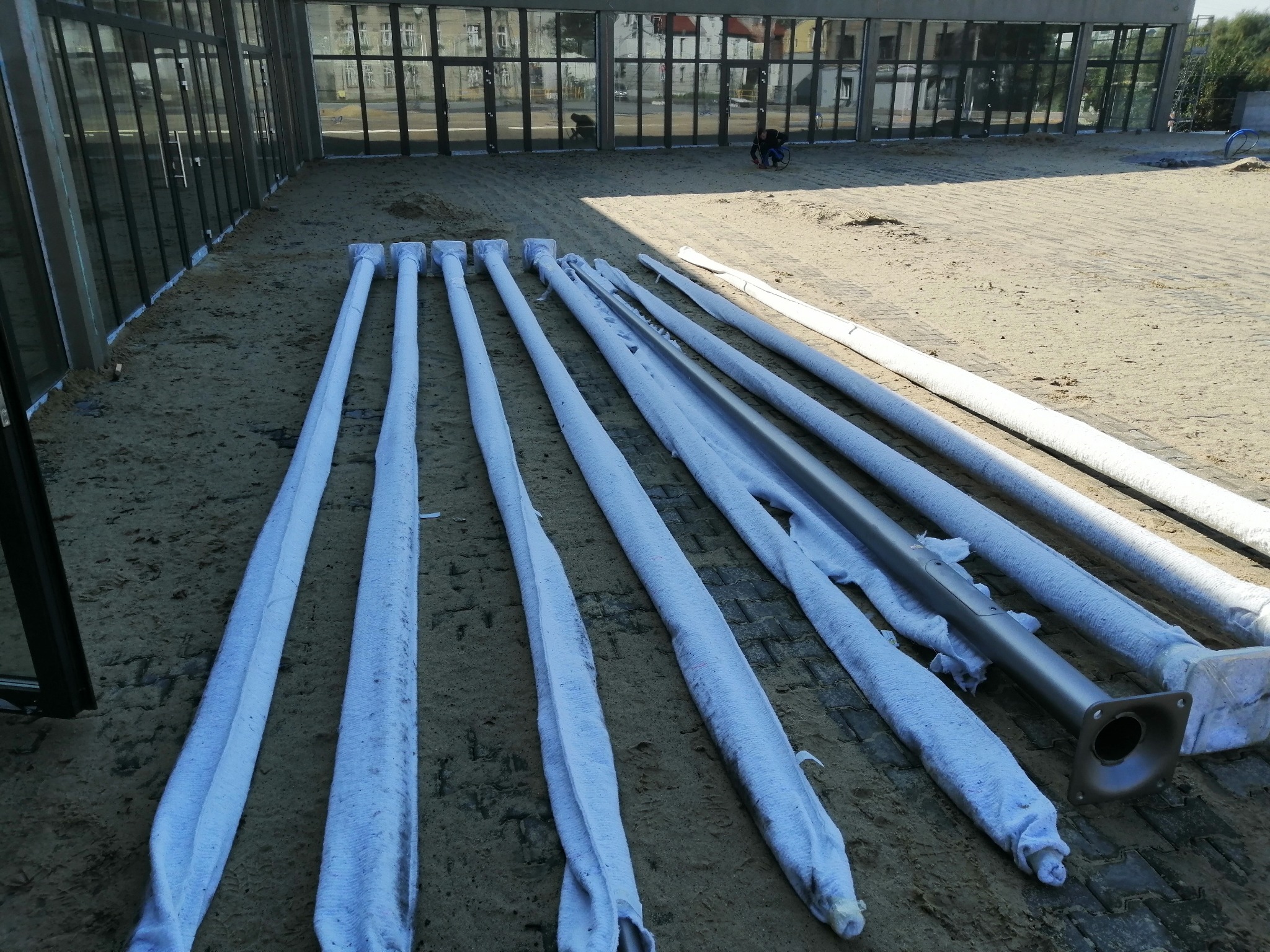 Przygotowane do montażu, długie, metalowe słupy oświetleniowe, częściowo owinięte ochronną folią, leżą na piaszczystym podłożu przed budynkiem z dużymi przeszkleniami. W tle widoczna sylwetka osoby...