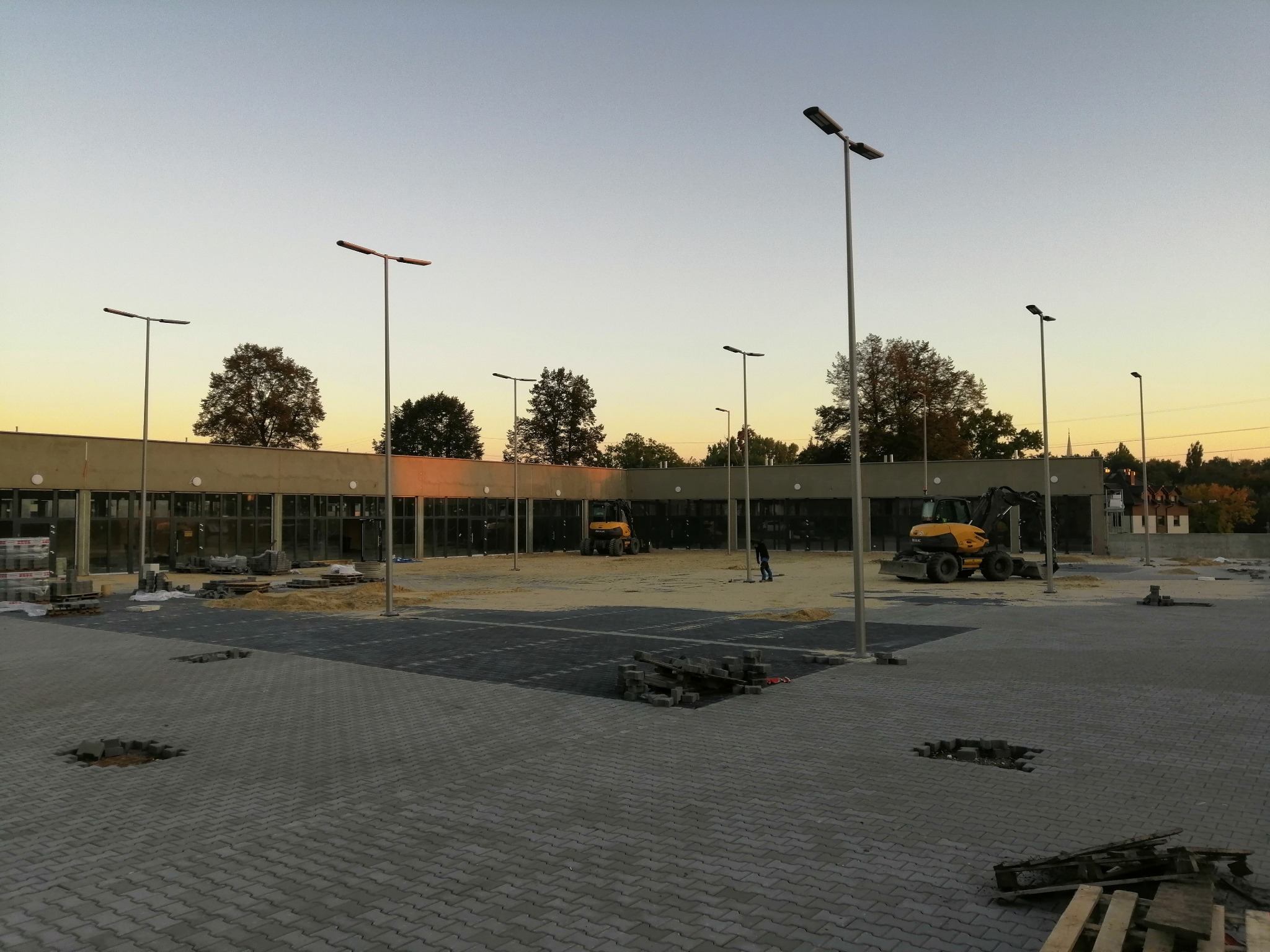 Plac budowy parkingu z kostki brukowej oświetlony latarniami, w tle budynek i koparki, widoczne palety z kostką i niedokończone fragmenty nawierzchni.