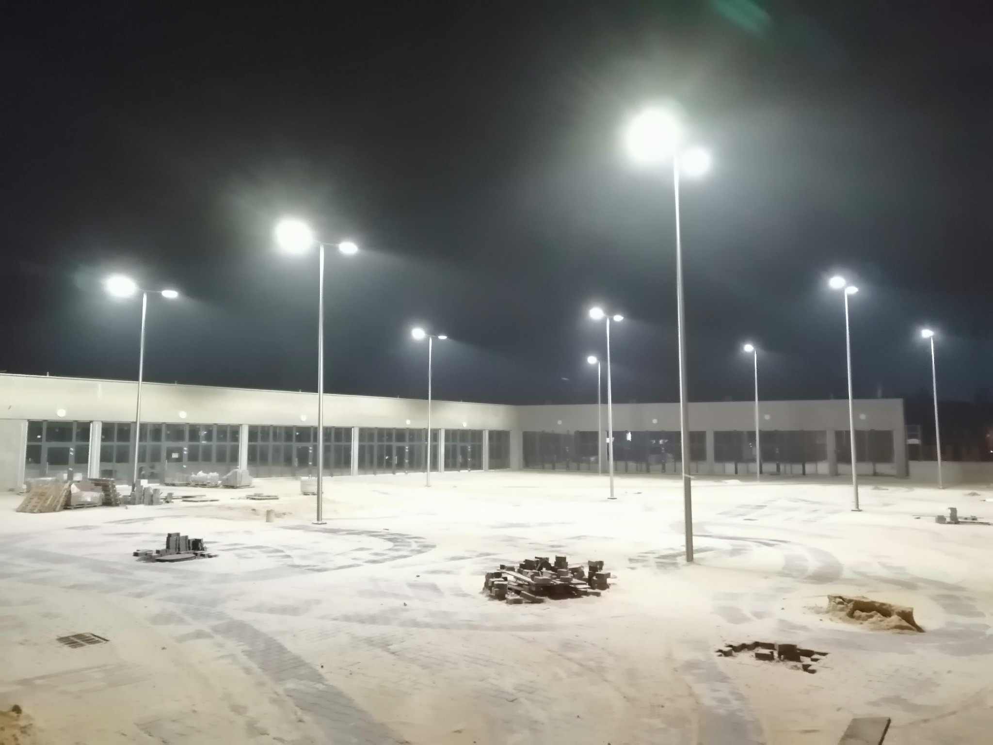 Plac budowy oświetlony nocą, widoczne lampy uliczne, materiały budowlane i fragment budynku z oknami.