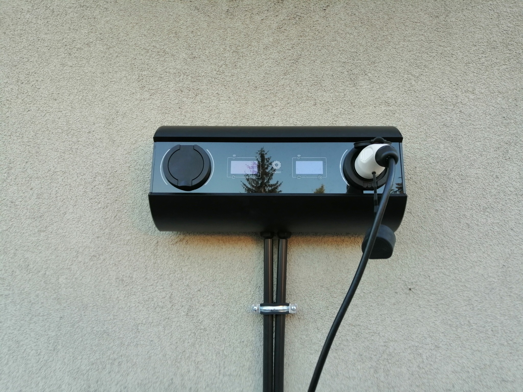 Czarna stacja ładowania pojazdów elektrycznych zamontowana na elewacji budynku, z widocznym kablem i wtyczką, dwa wyświetlacze i odblask drzewa w panelu.